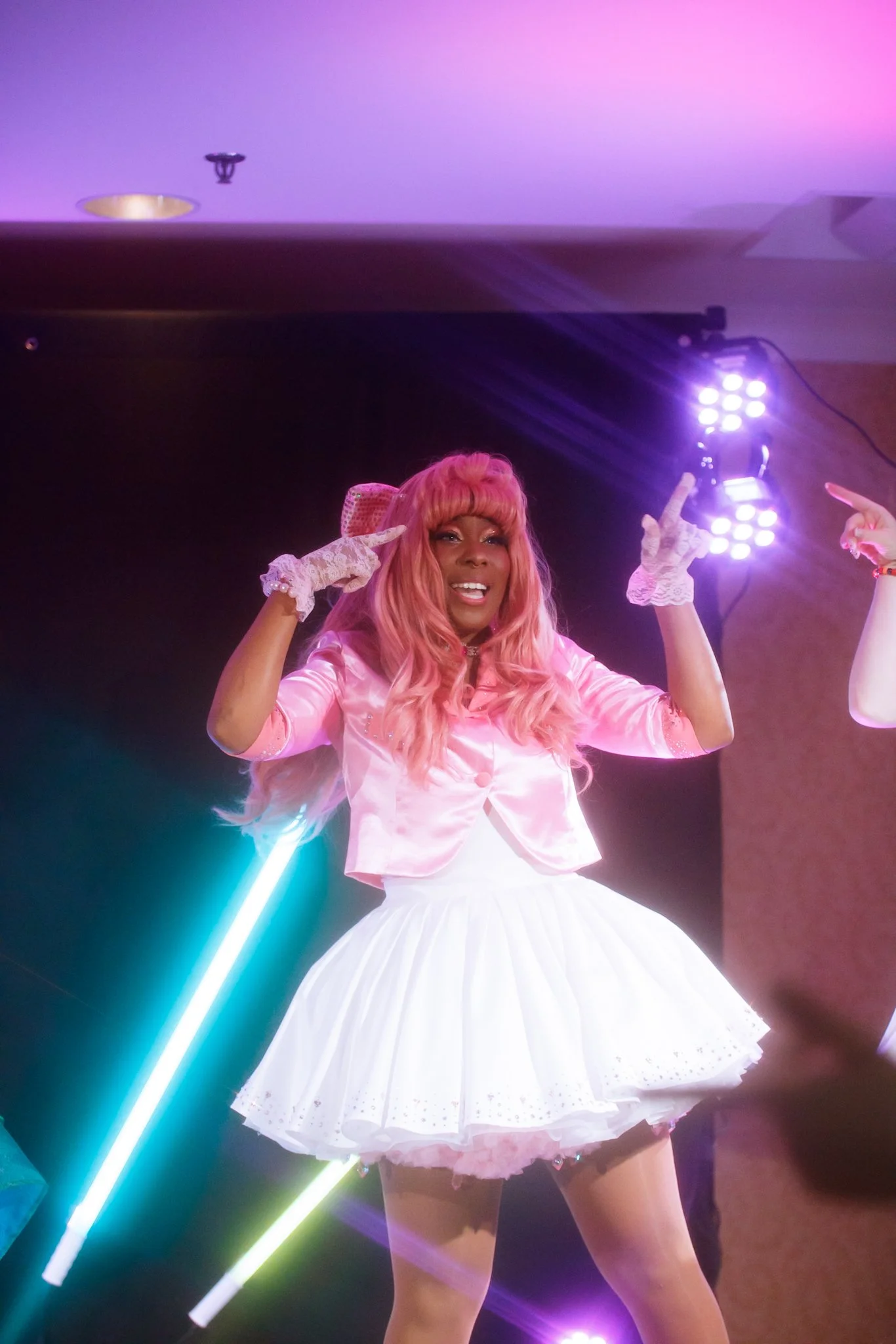 Performer with pink hair and pink satin jacket, white skirt, and lace gloves on stage with colorful lights