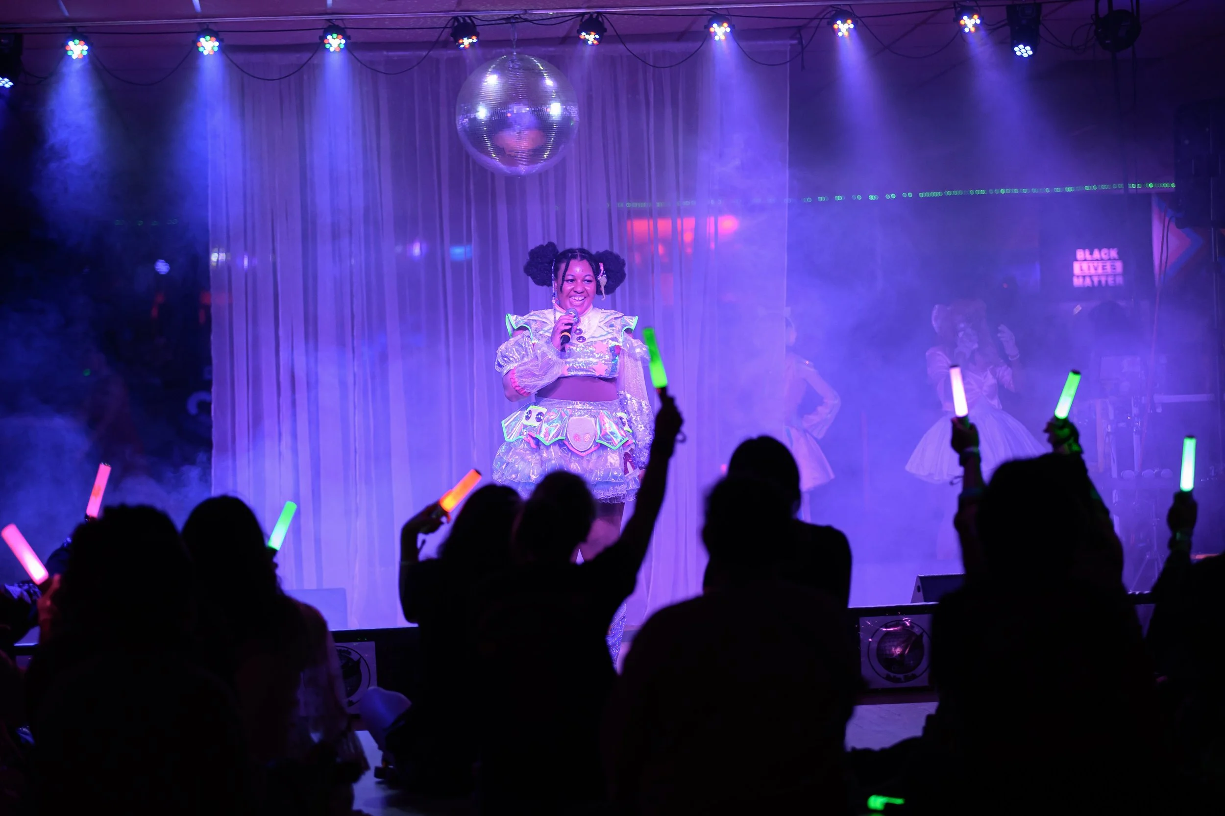 A performer smiling on stage, wearing a shiny, colorful costume, with children in the audience waving glow sticks in a dark, vibrant club setting with purple lighting and a disco ball overhead.