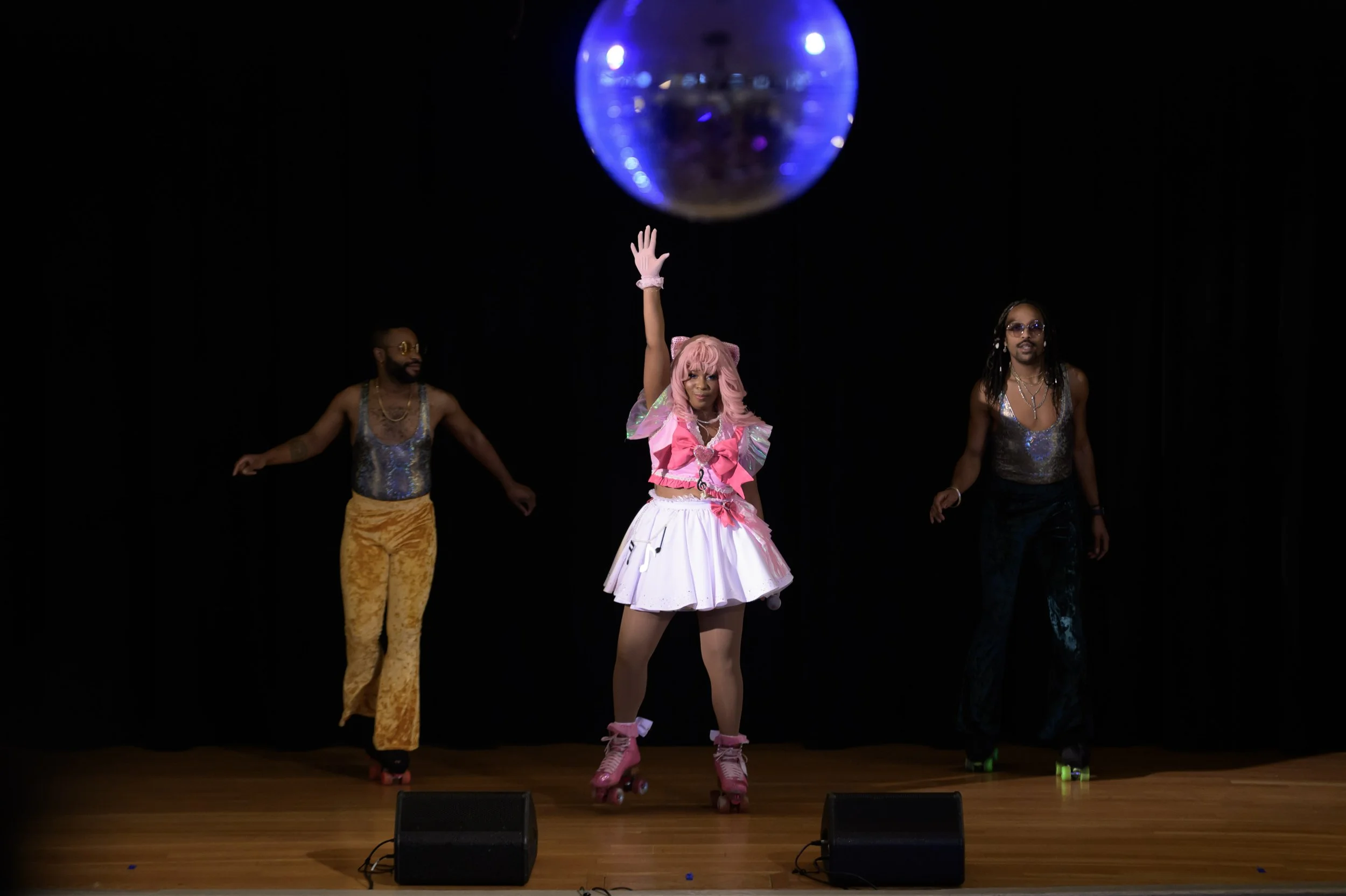 Three performers on stage; a woman in the center with pink hair, pink roller skates, and a colorful outfit, raising her hand; two men on either side in shiny tops and bell-bottom pants.