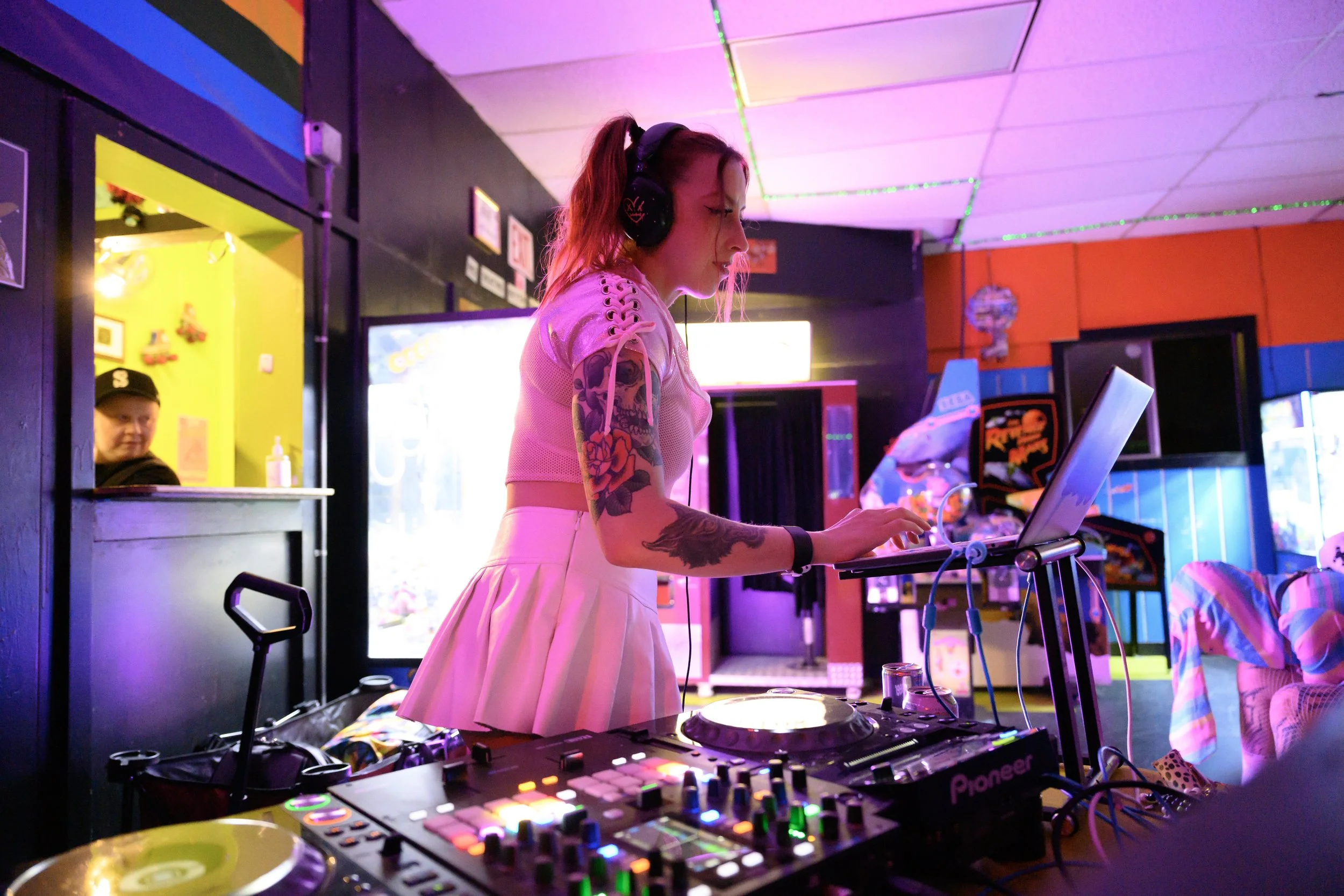 A female DJ with tattoos on her arm, wearing headphones, is focused on her laptop and DJ equipment in a colorful, club-like setting with neon lighting and arcade machines in the background.