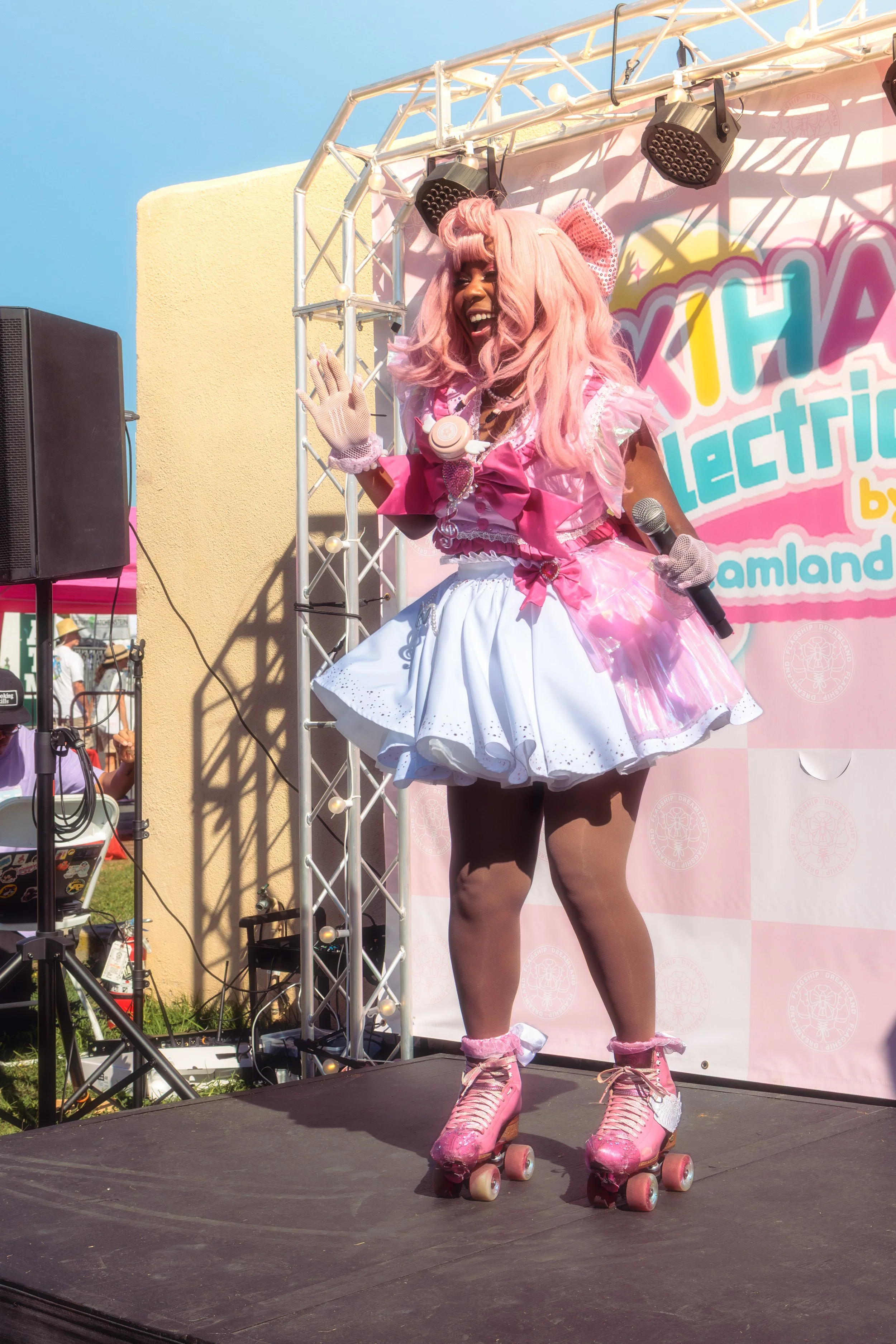 A performer in pink and white roller skates, a pink wig, and a pink and white outfit, smiling and waving on stage at an outdoor event with a pink and blue banner in the background.