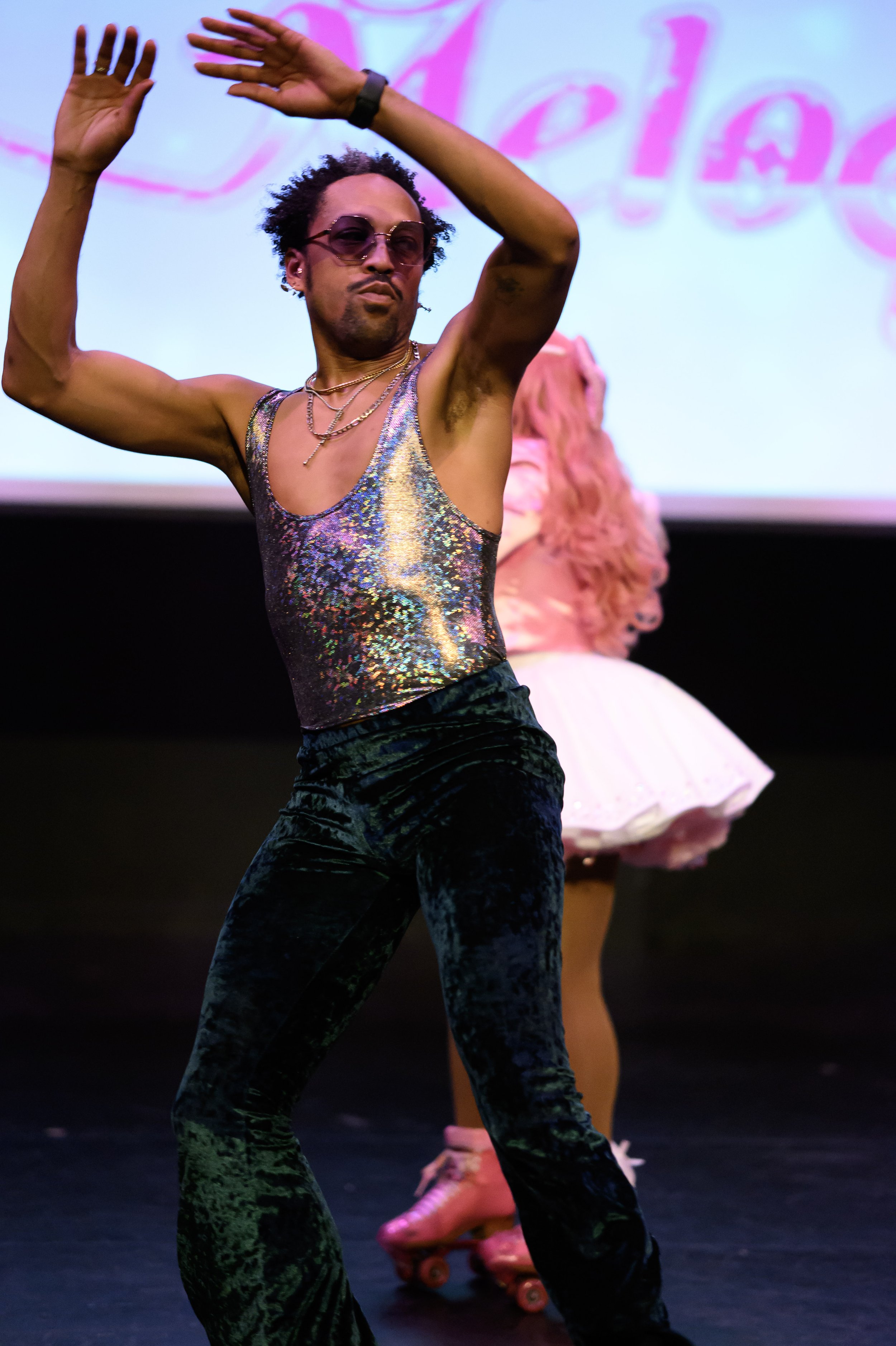A person dancing on stage wearing a shiny, iridescent tank top, dark velvet pants, sunglasses, and layered necklaces, with a woman on roller skates in the background.
