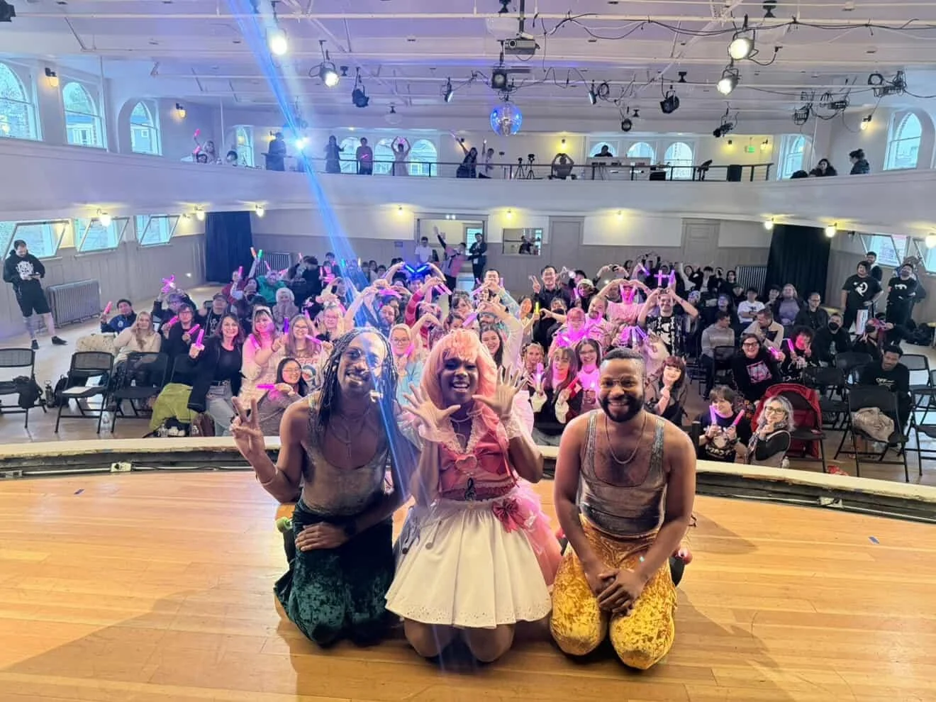 People on stage and in audience at a lively event in a large auditorium with colorful lighting, some holding glow sticks, and performers taking a photo at the front.