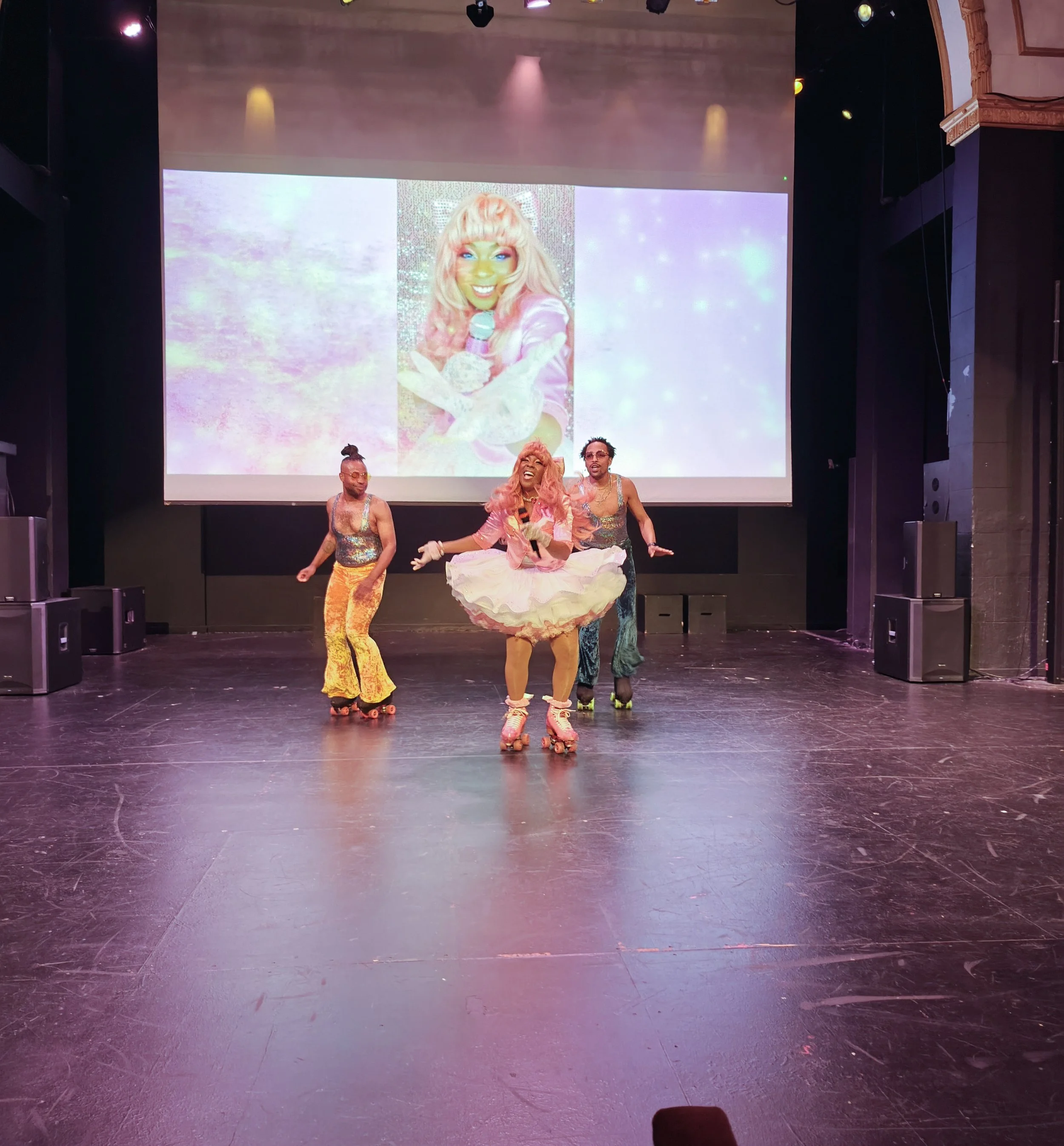 Three performers roller skating on stage with colorful costumes, performing in front of a large screen displaying a woman's digital portrait. The stage has a dark floor and surround sound speakers.
