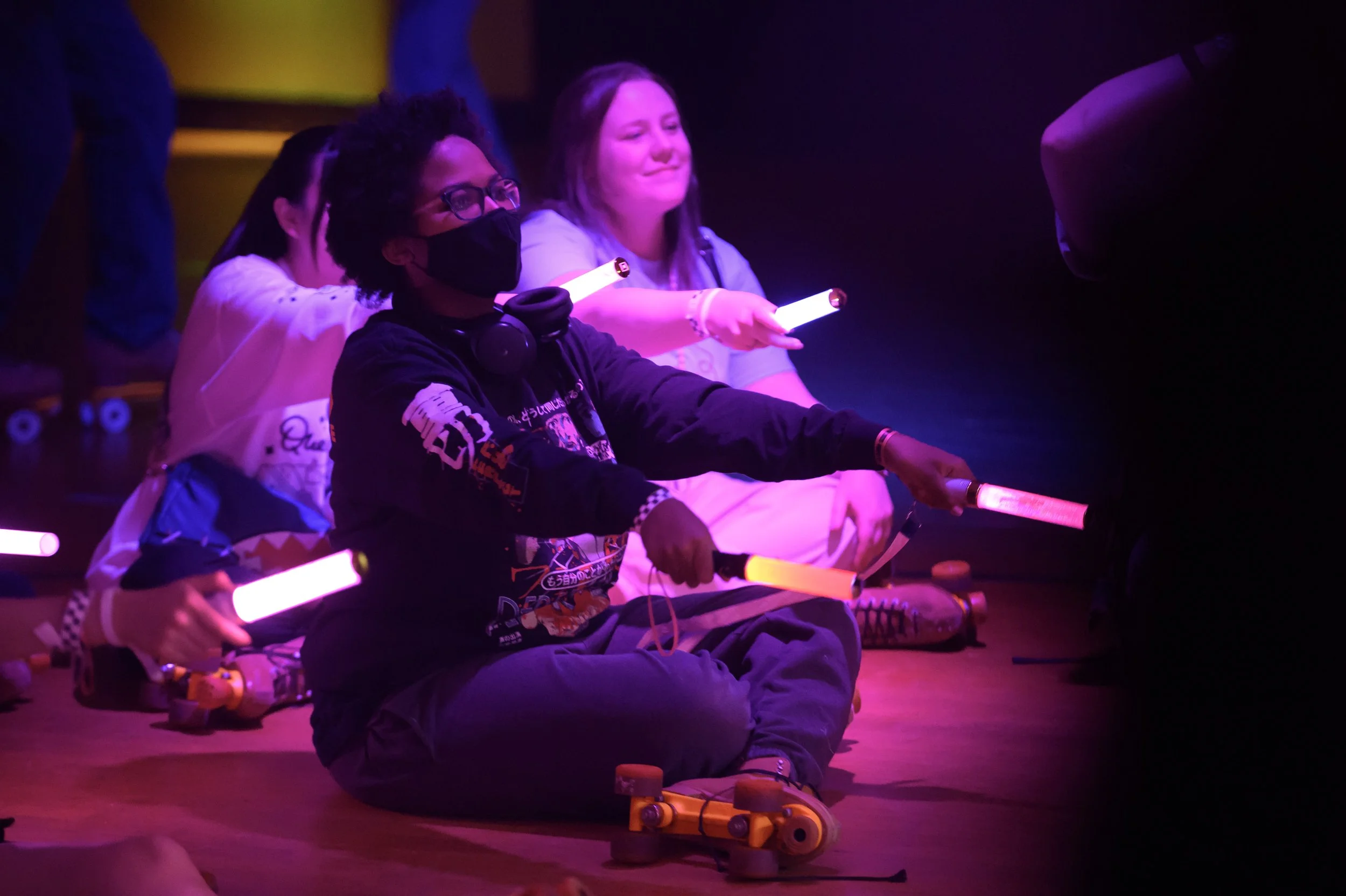 People sitting on the floor in a dark room, illuminated by colorful lights, holding glowing light sticks, with some wearing masks.