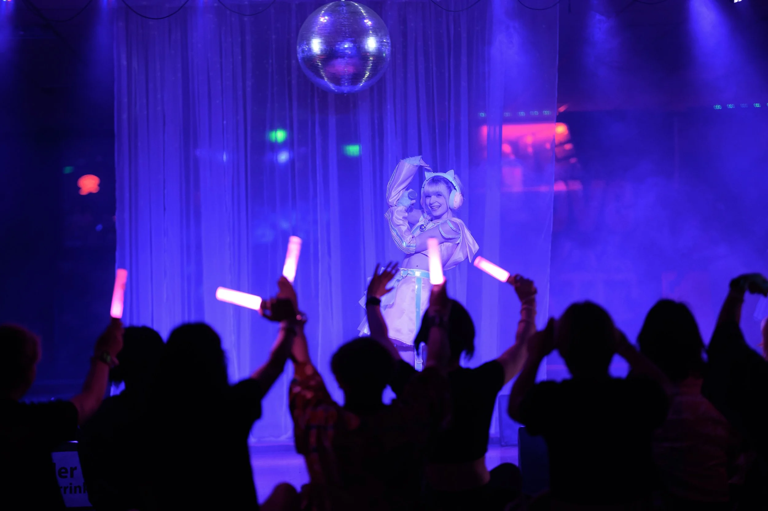 A woman dressed in a pastel outfit with sunglasses and headphones on stage, smiling, with a disco ball overhead and a purple curtain backdrop. Audience members are seated and holding glow sticks.