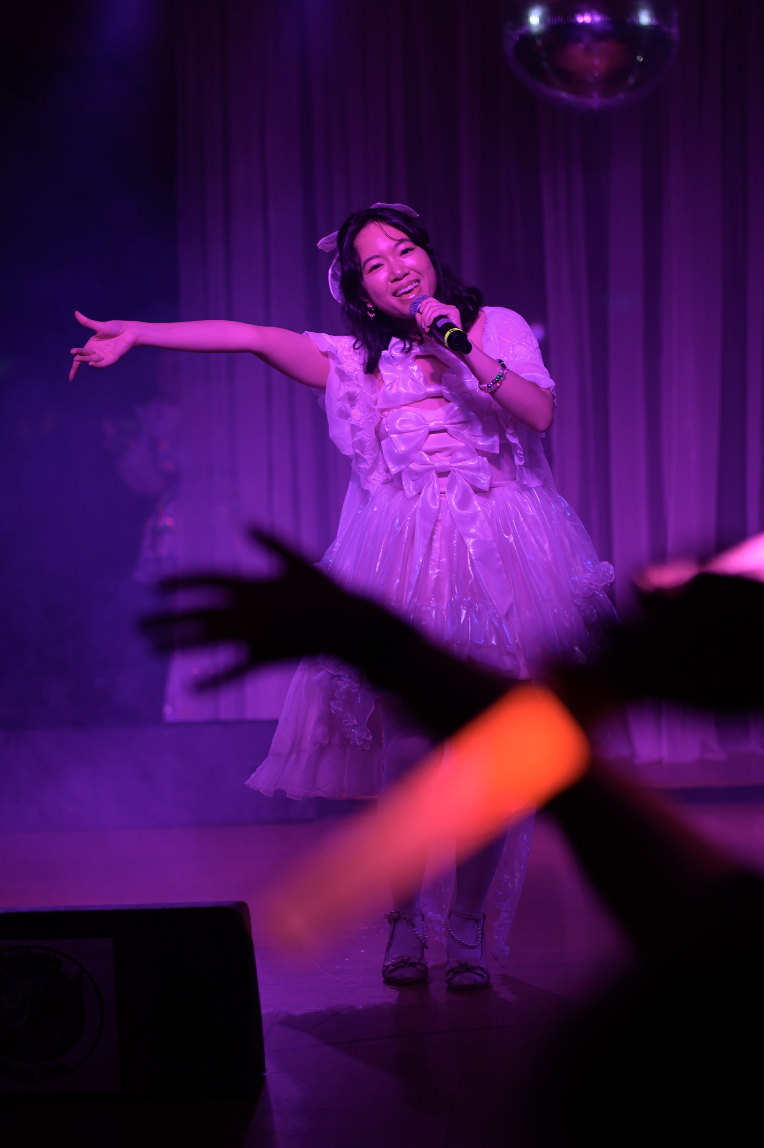 A woman performing on stage with purple lighting, wearing a pink dress, holding a microphone, smiling, with an audience member's hand in the foreground.