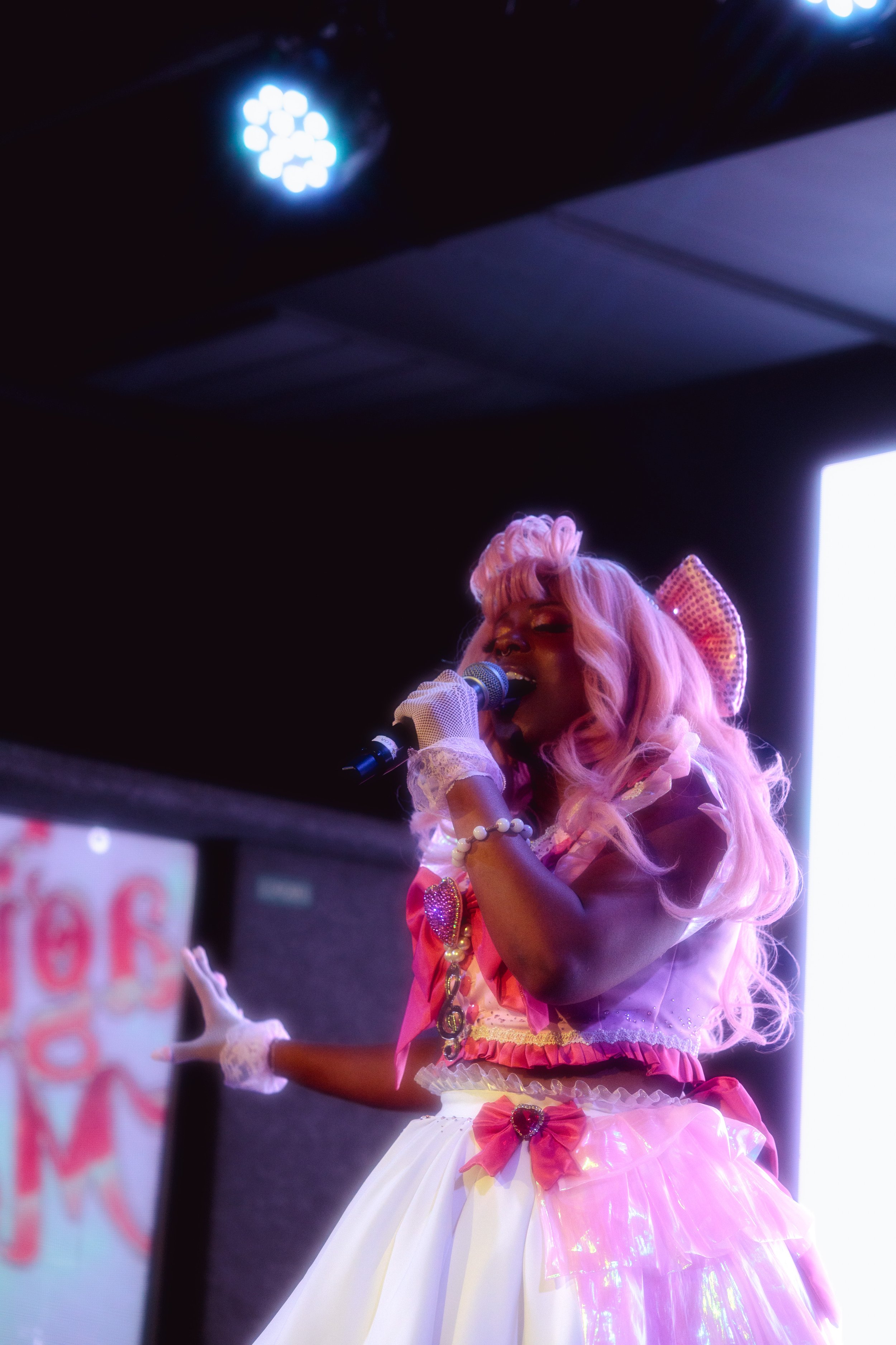 Performer singing into a microphone on stage, wearing a pink and white dress with bows and accessories, with pink hair styled in curls.
