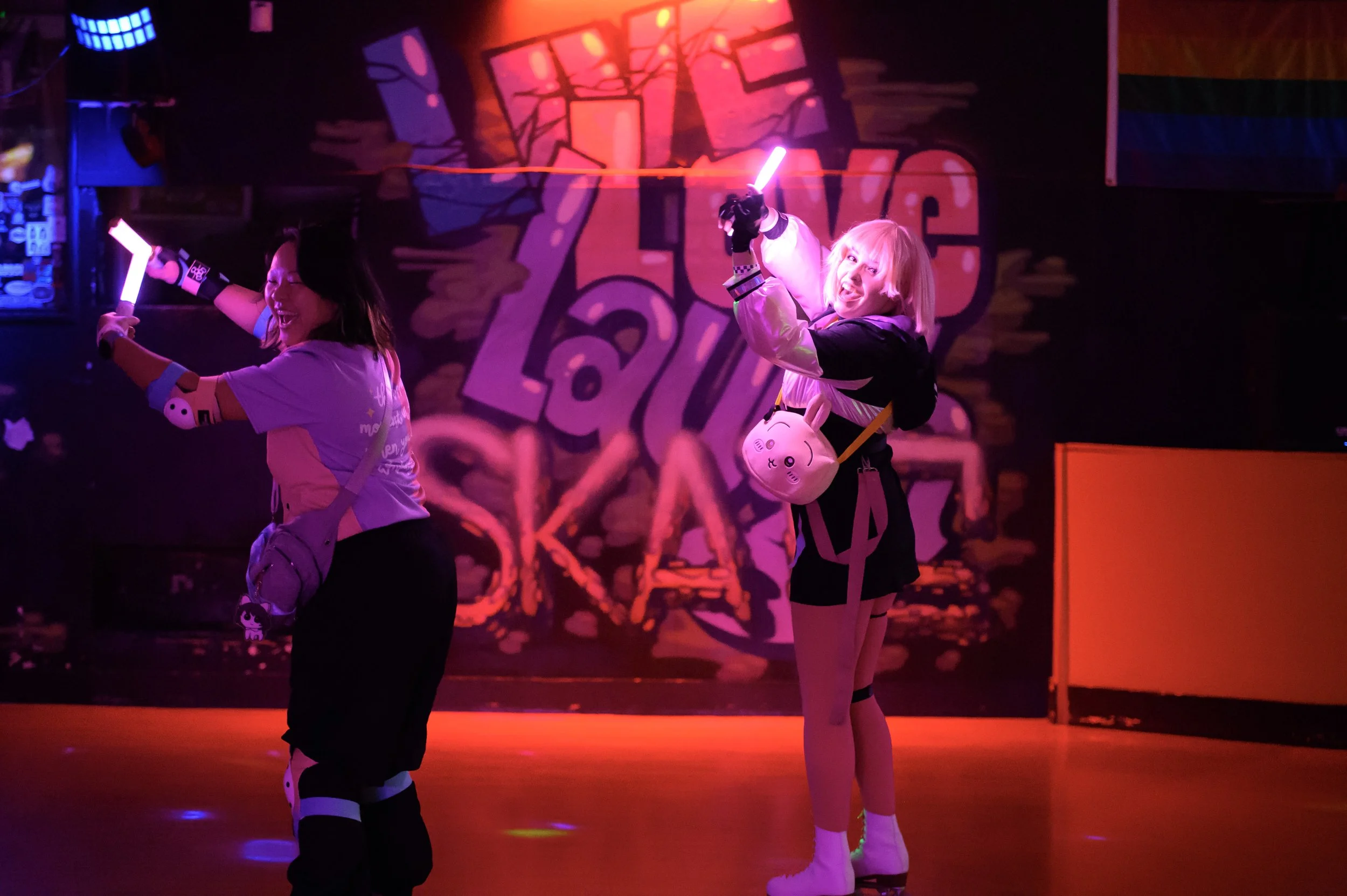 Two women with light-colored hair and playful expressions roller skating in a neon-lit indoor skate rink. The woman on the right is holding a glowing pink light stick and wearing a small plush bag with a cute character face. The background features v
