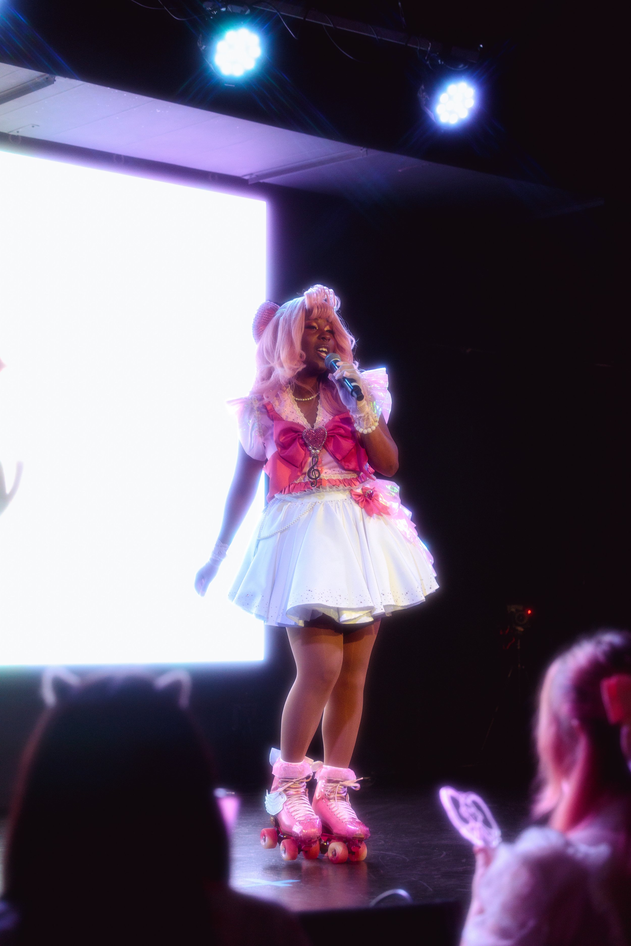 Person dressed in pink and white costume with roller skates performing on stage, holding a microphone, with audience members visible in foreground.