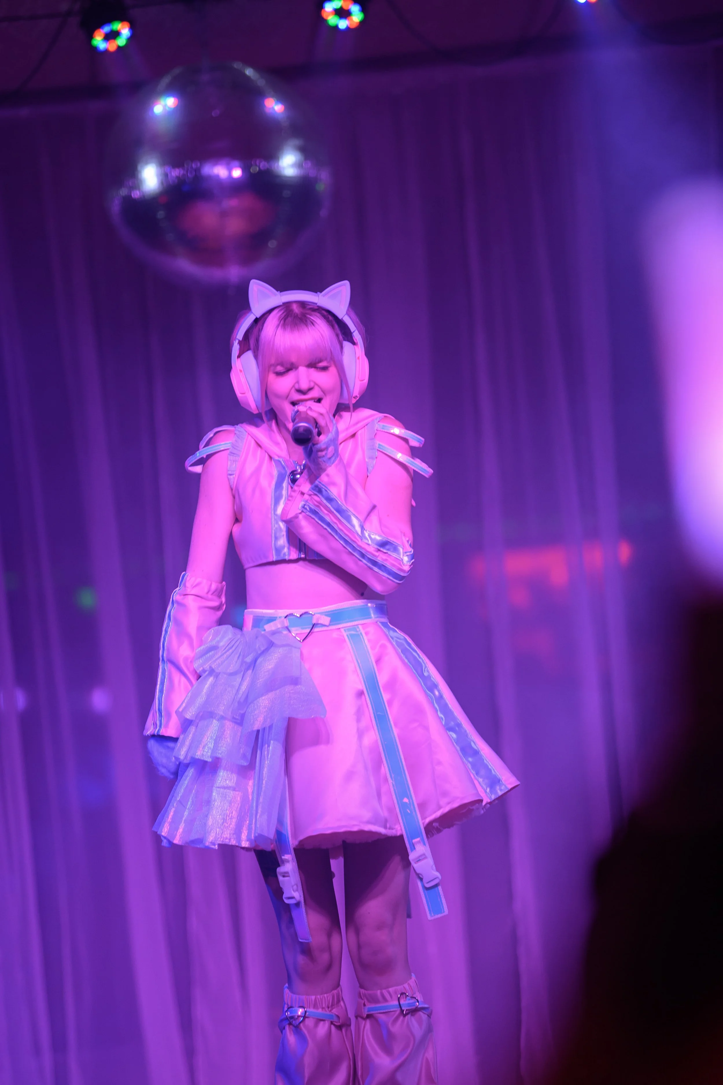 A young woman dressed in a colorful, futuristic costume with a pink skirt, matching top, and cat ear headband, singing into a microphone on stage under purple lighting with a disco ball overhead.