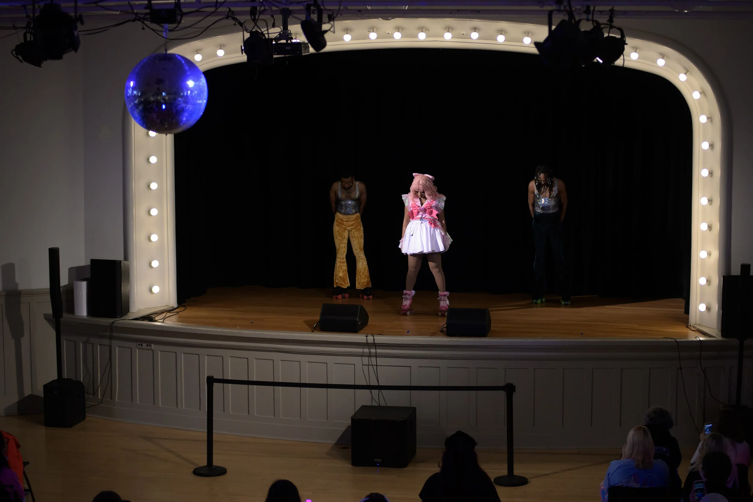 Three performers on a stage, with one dressed in a pink and white costume with roller skates, and two others in colorful outfits, under stage lights and a blue disco ball.