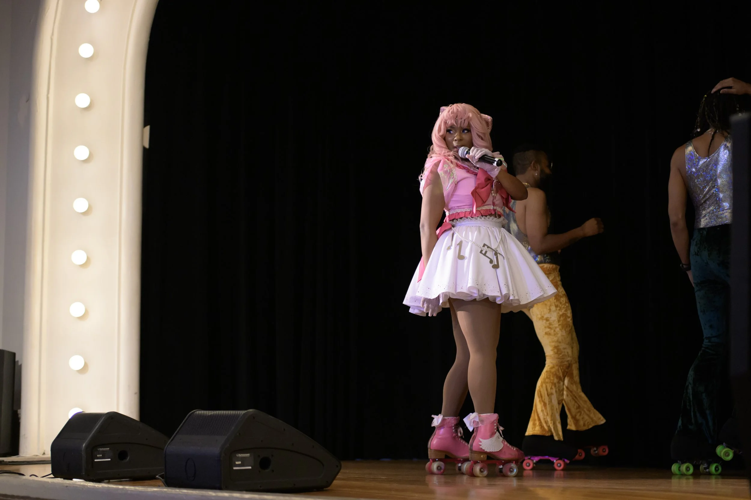 Performer wearing a pink costume with a tutu skirt and pink roller skates, holding a microphone on stage during a dance or performance event.