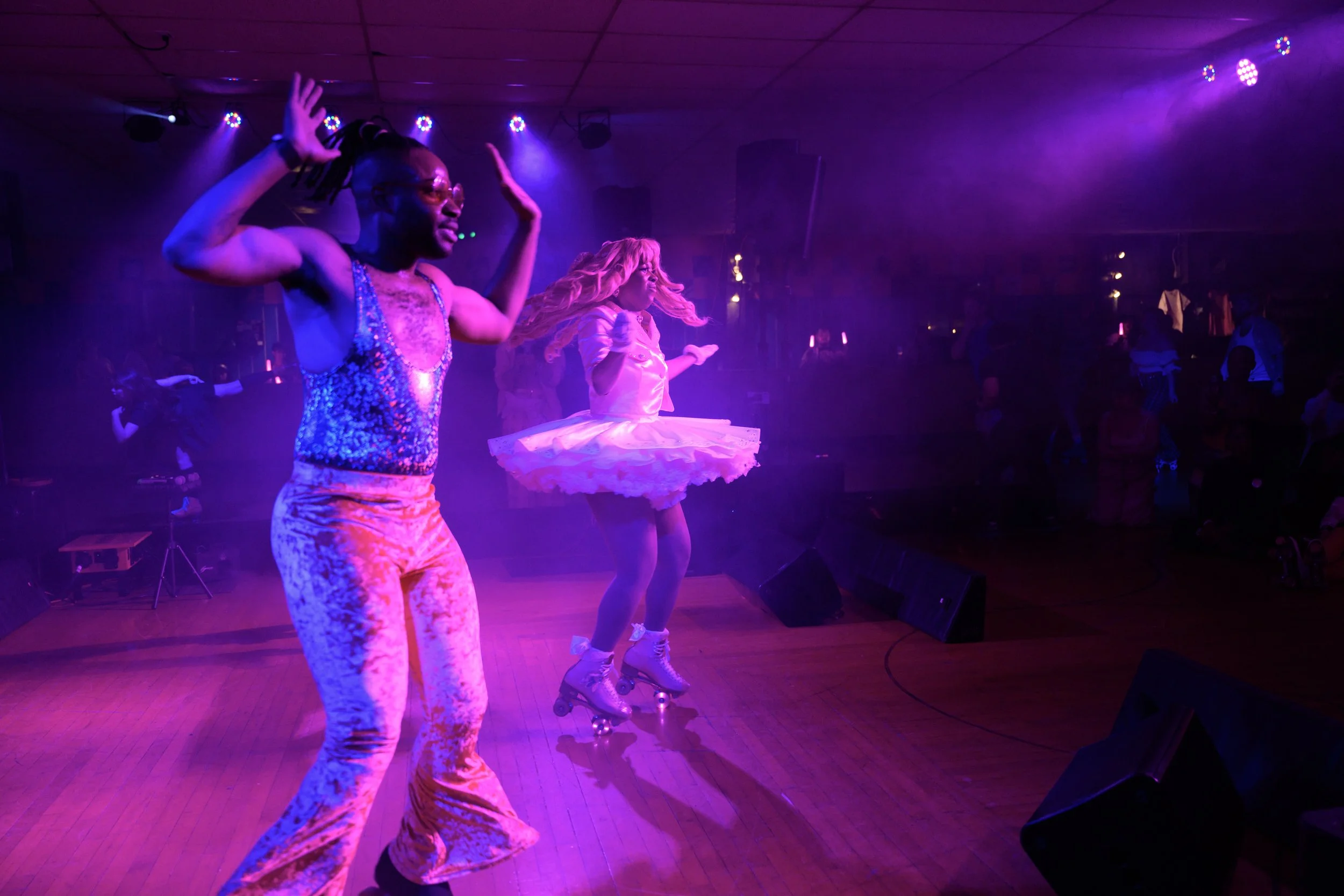 People dancing on a stage under purple and pink lights, one in colorful retro clothing, another in a pink ballet tutu and roller skates.