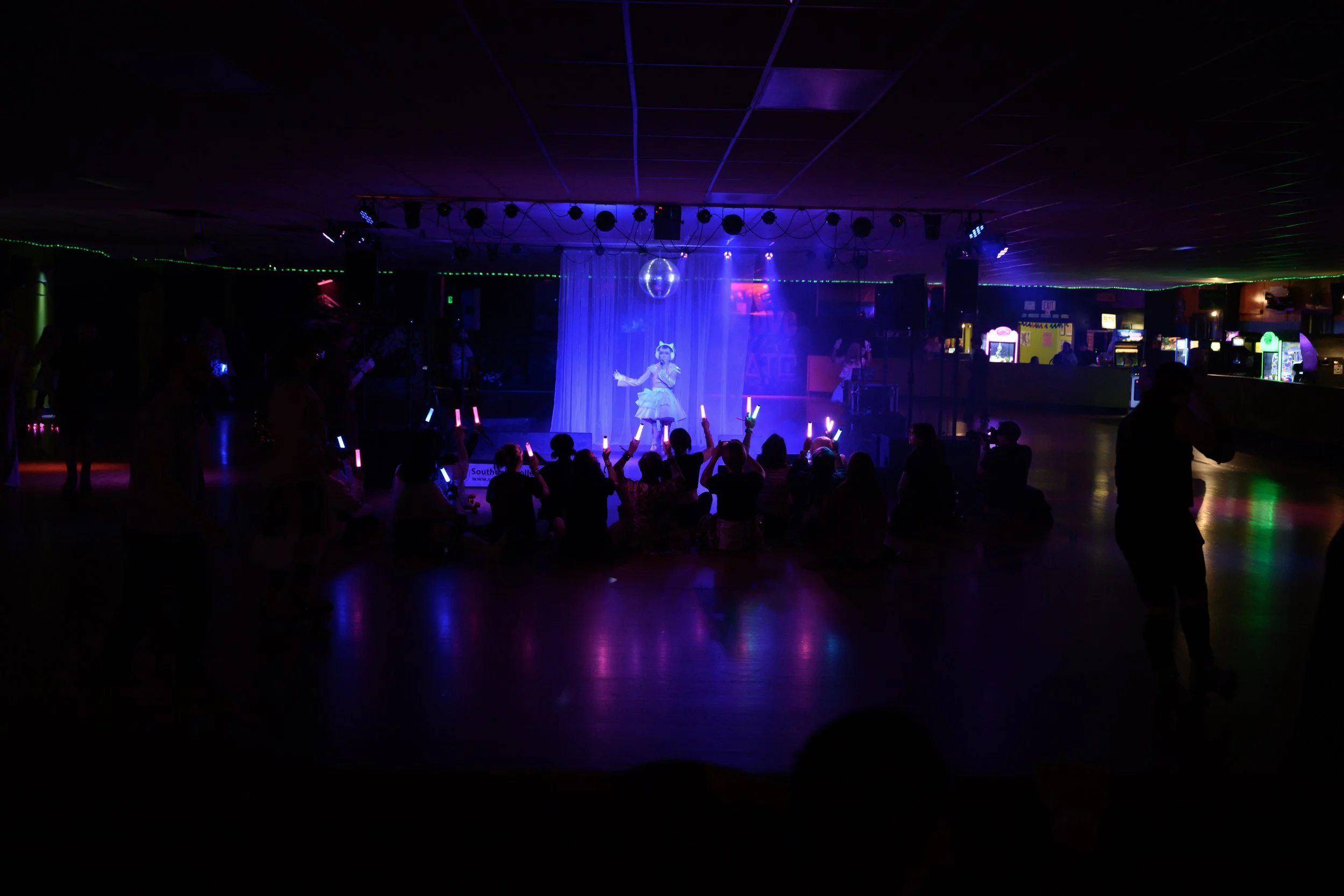 A performer on the rink floor, with people sitting on the floor holding light sticks, in a dimly lit room with colorful neon lights and arcade machines in the background.