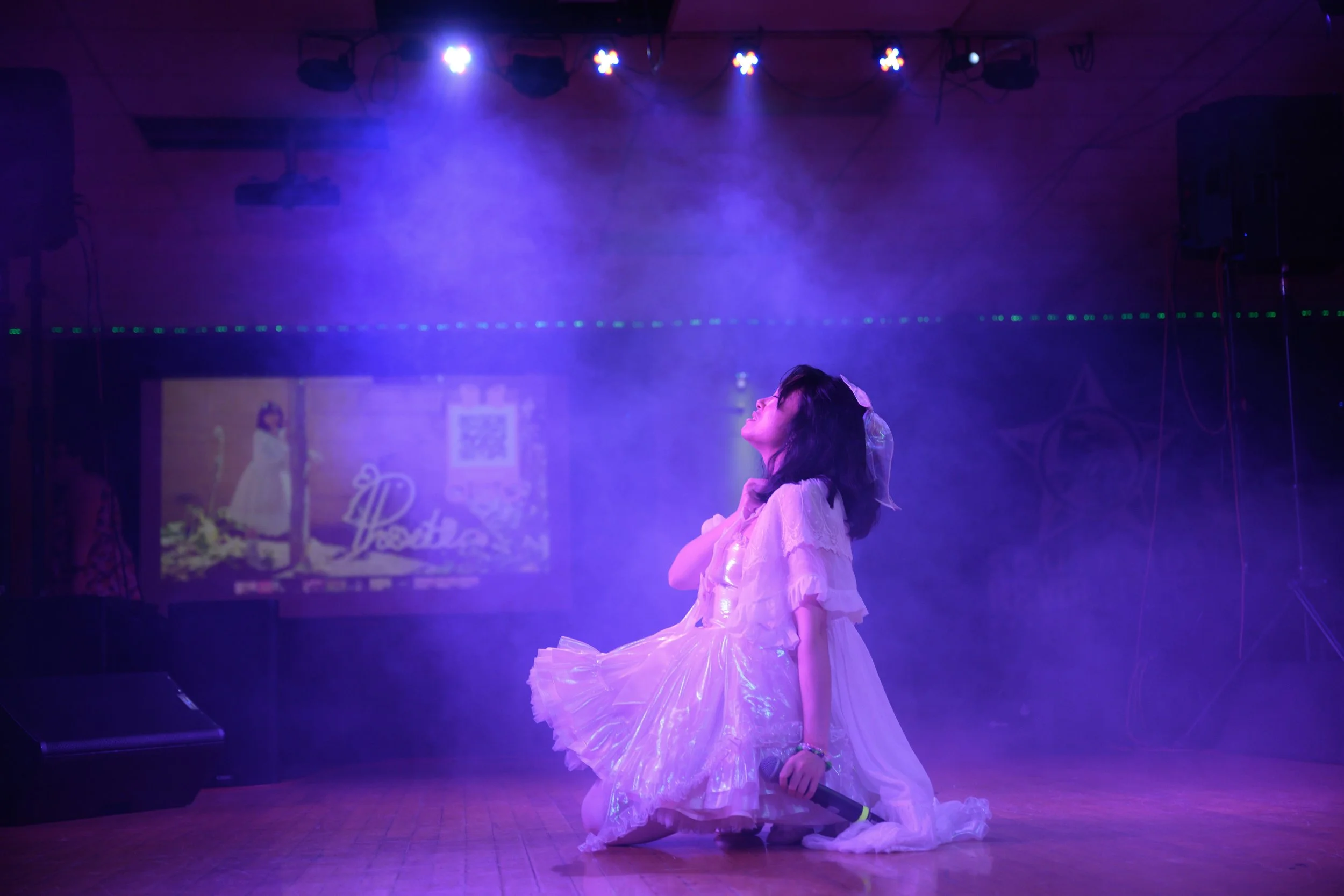 A young girl dressed in a white, frilly dress performs on stage. She kneels with a microphone in her hand, looking up with her eyes closed. Stage lights and fog create a purple ambiance, and a screen in the background displays a woman and some text.