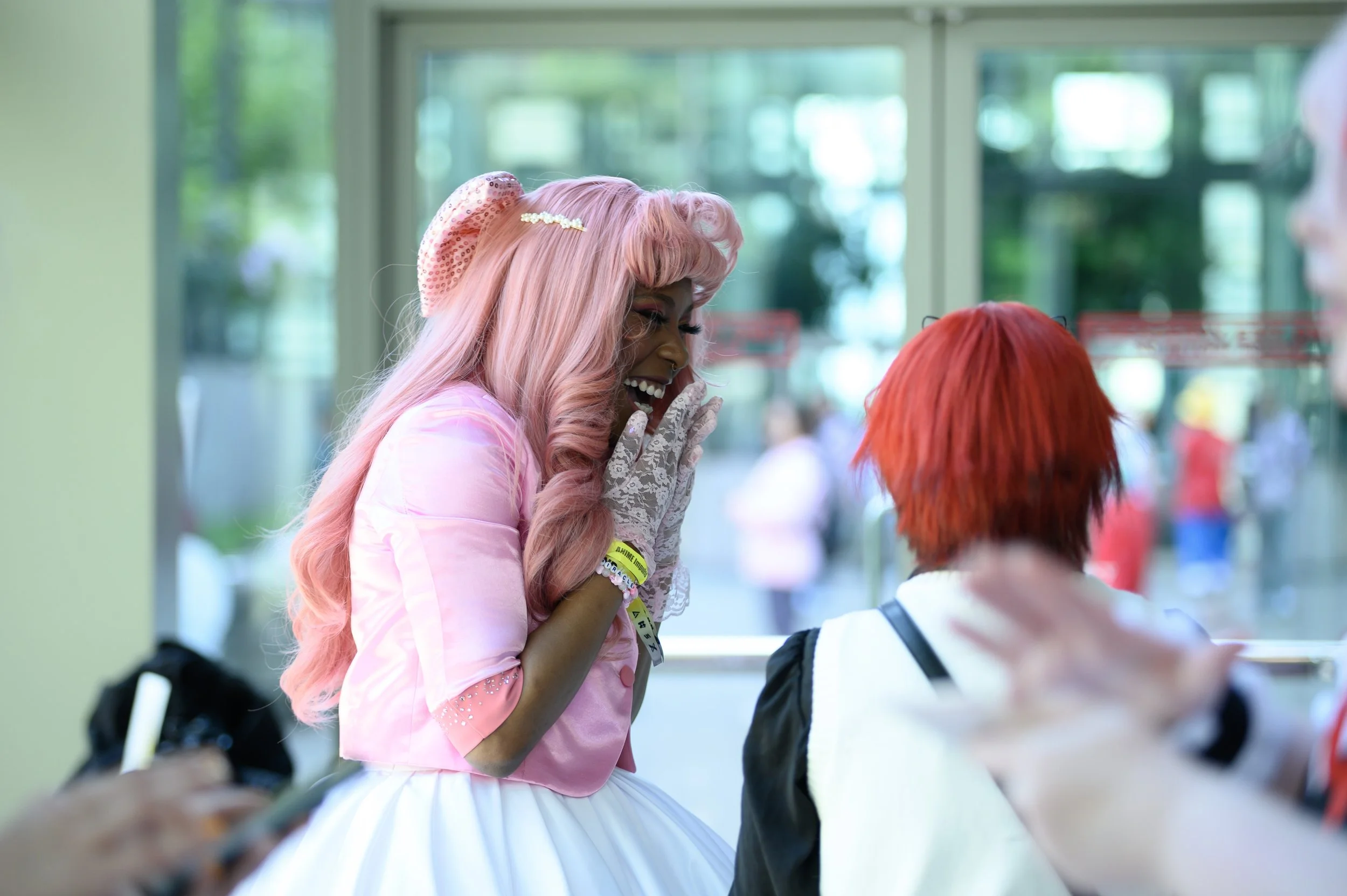Two women with pink and red hair, dressed in pastel and black clothing, are engaging in a lively conversation indoors with a large window behind them showing an outdoor scene with blurred people.