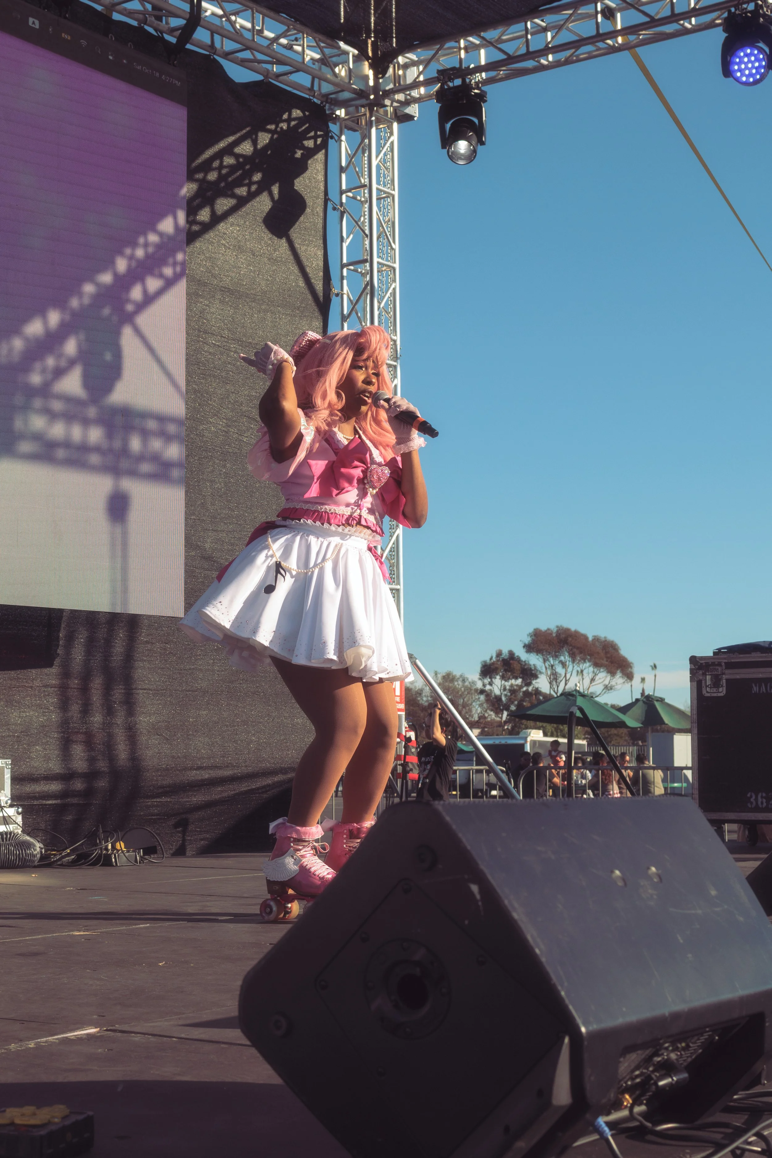 Performer in a pink and white costume with pink wig and roller skates singing on stage during an outdoor event.