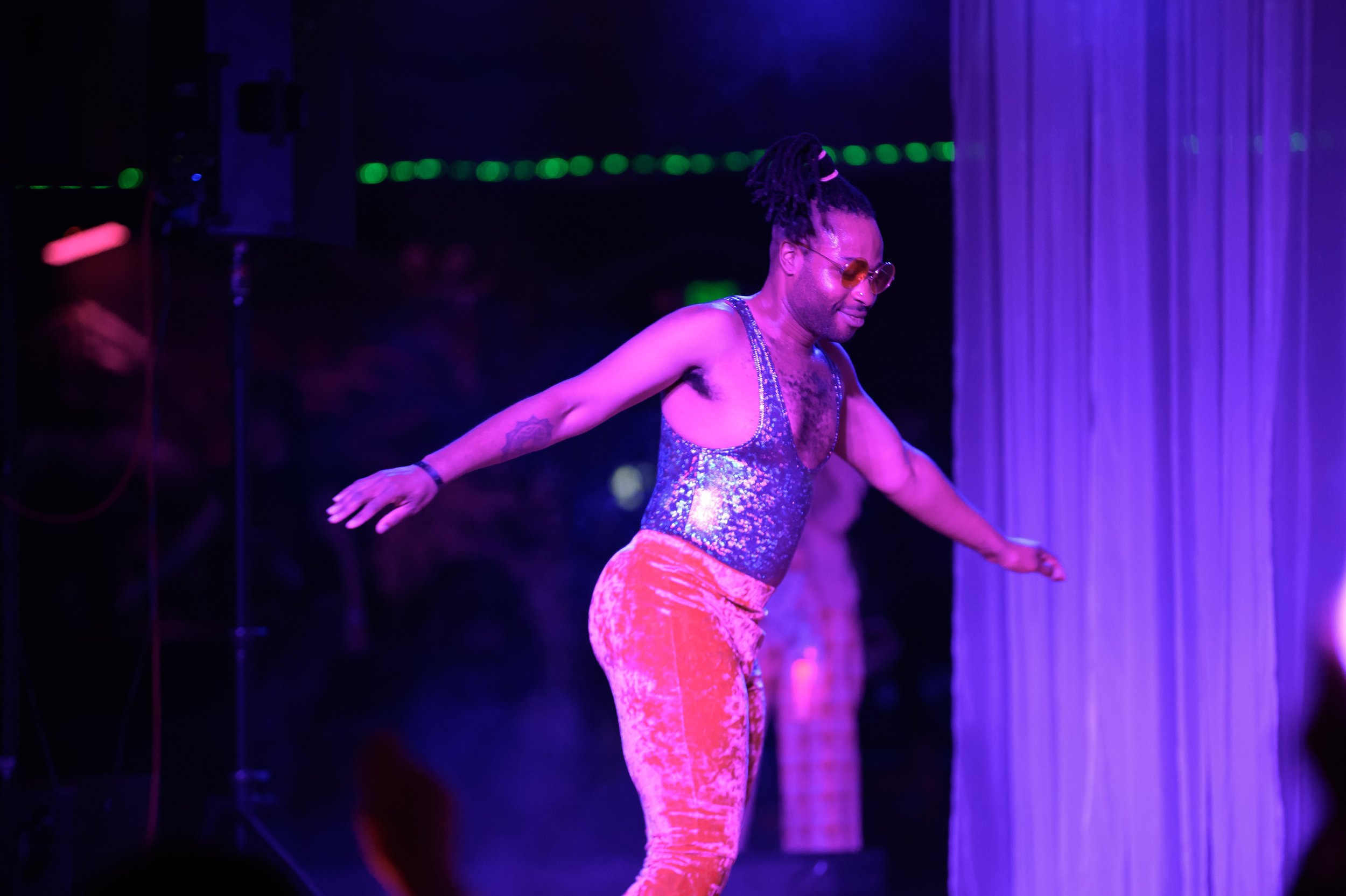 A man with dreadlocks wearing sunglasses, a shiny silver tank top, and pink pants stands with arms outstretched on stage under purple lighting.
