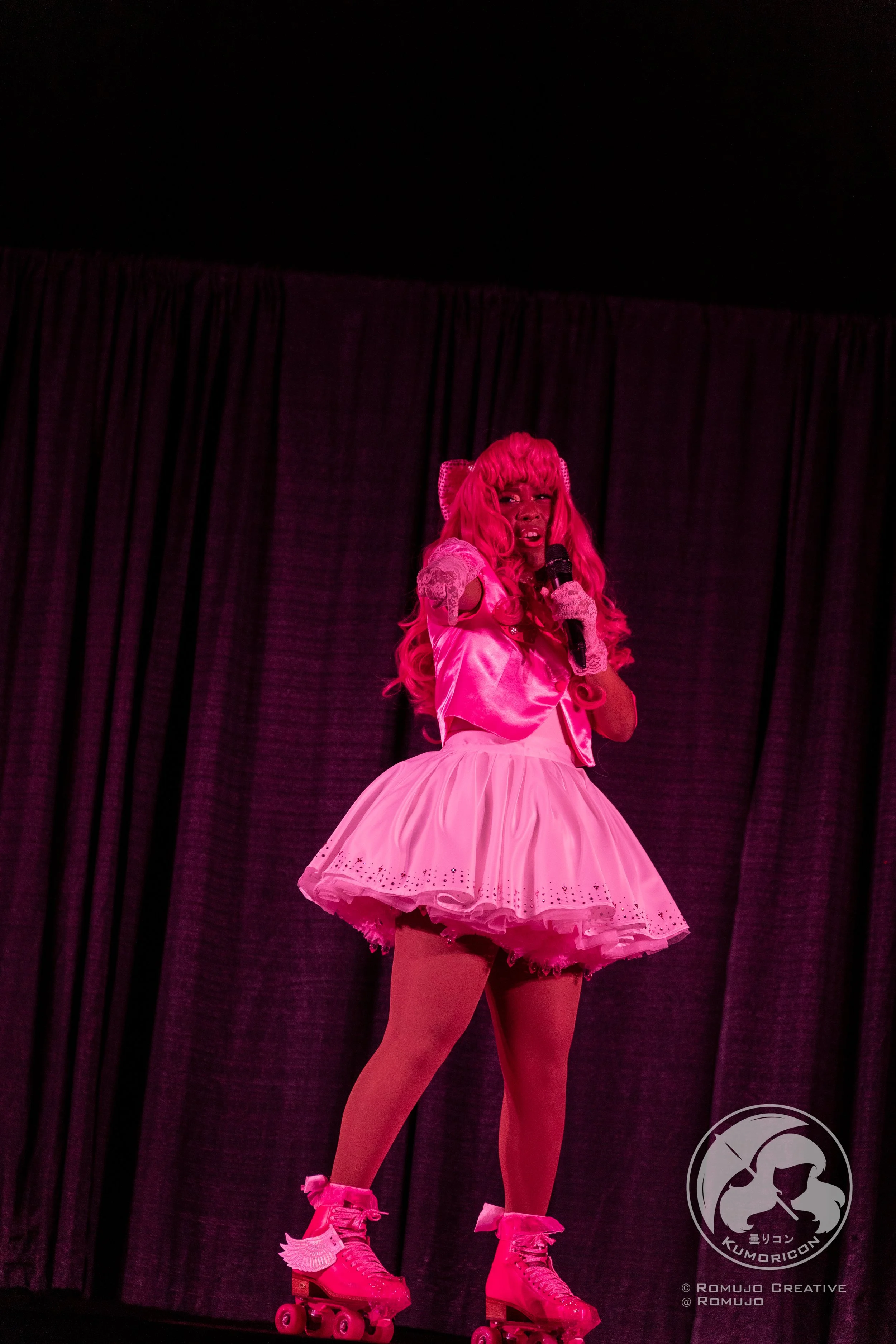 Person dressed in pink, wearing a pink wig, pink dress, and pink roller skates, performing on stage with curtains in the background.