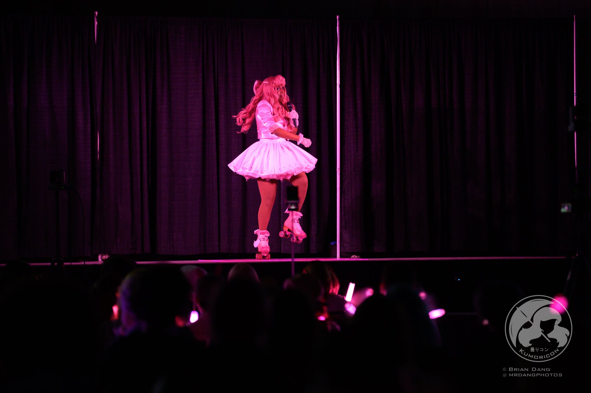 Performer dressed in pink cosplay costume on stage with a microphone, roller skates, and pink wig, performing under purple stage lighting with black curtains in the background.