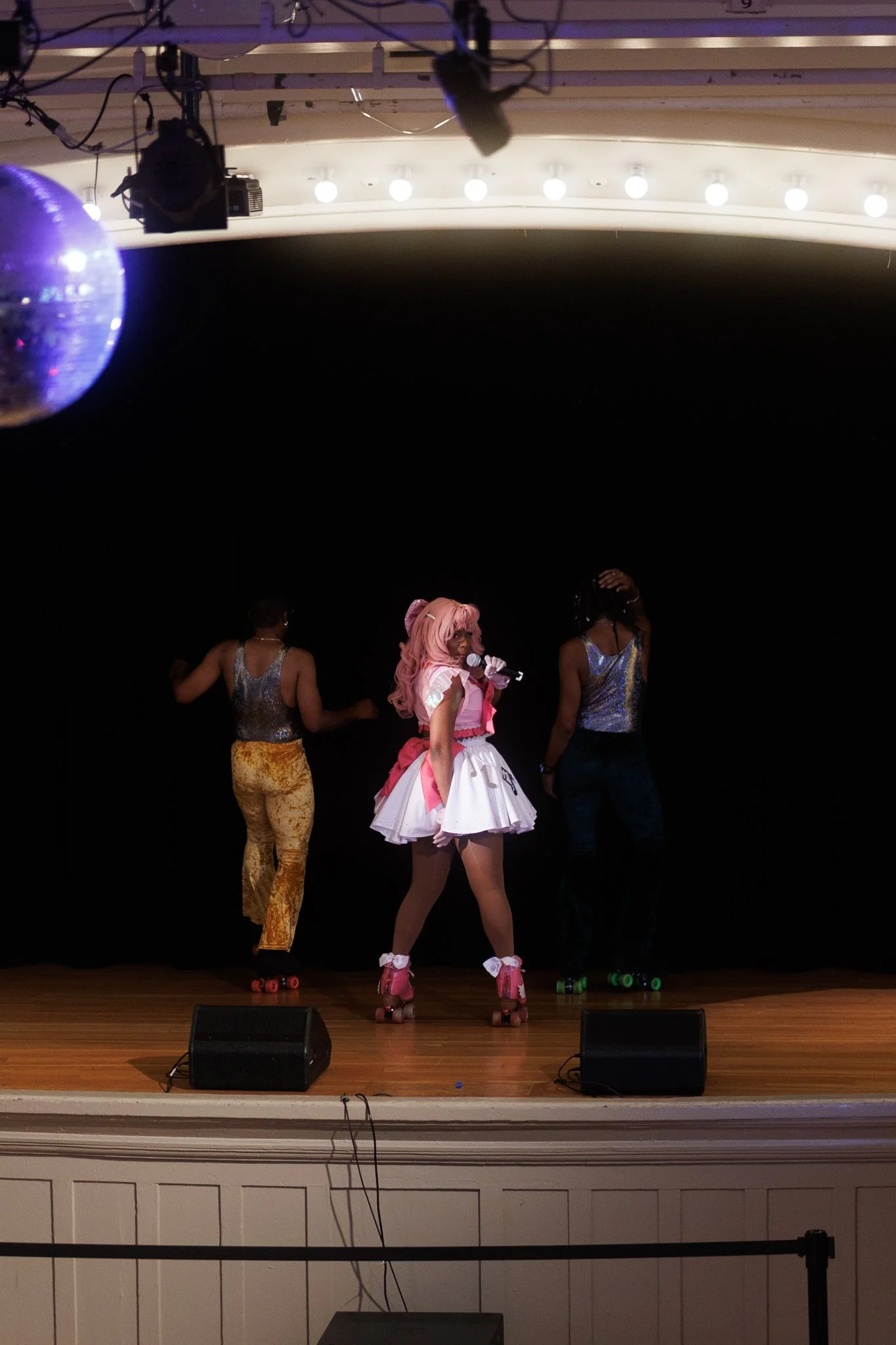 A person dressed as a performer with pink hair and a pink and white costume on roller skates on stage, holding a microphone, with two dancers in colorful outfits behind them, in front of a black backdrop on a stage with audio equipment.
