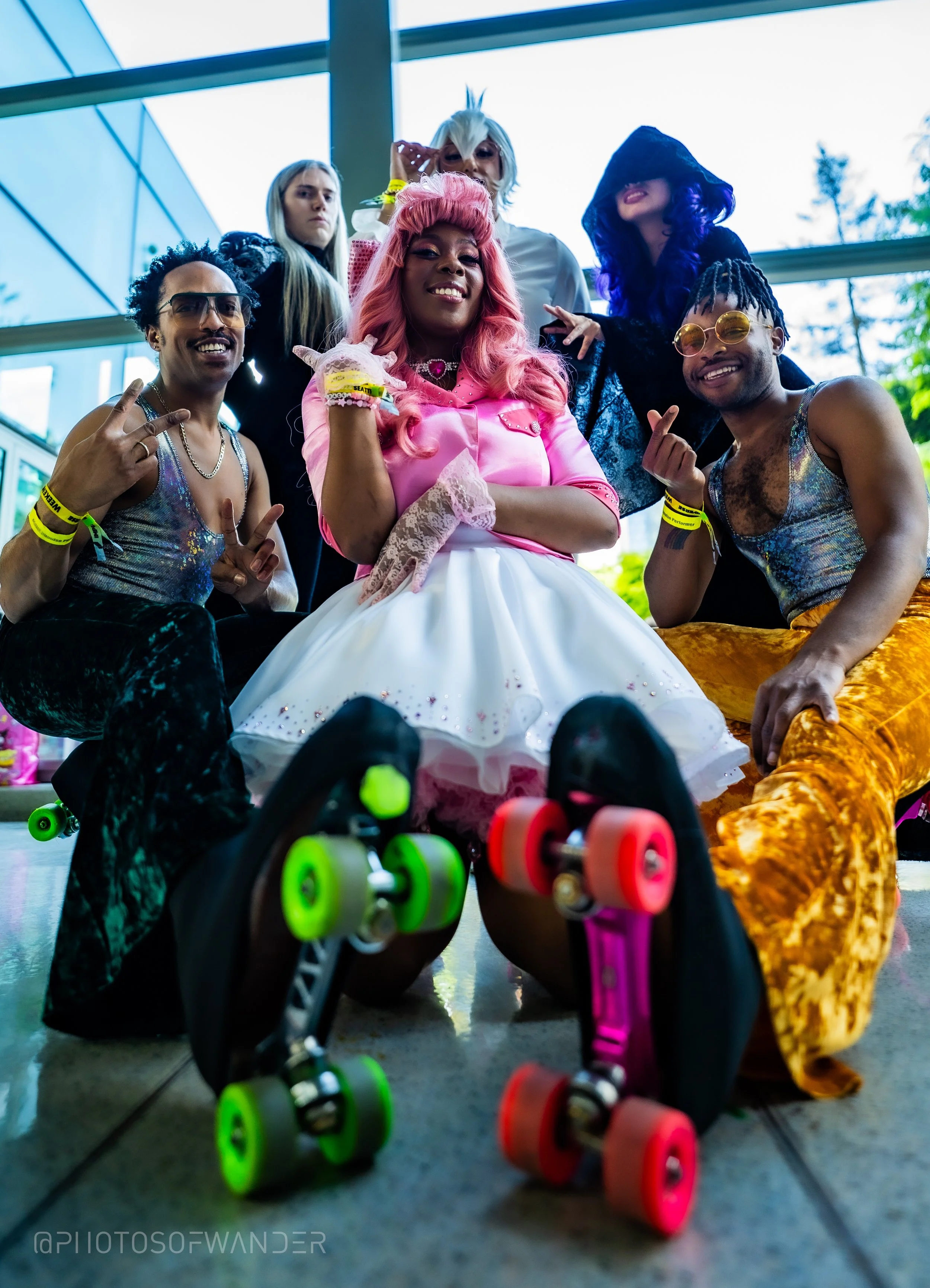 Group of six young people dressed in colorful, eclectic clothing posing for a photo indoors with large windows, with skateboards on the floor in front of them.