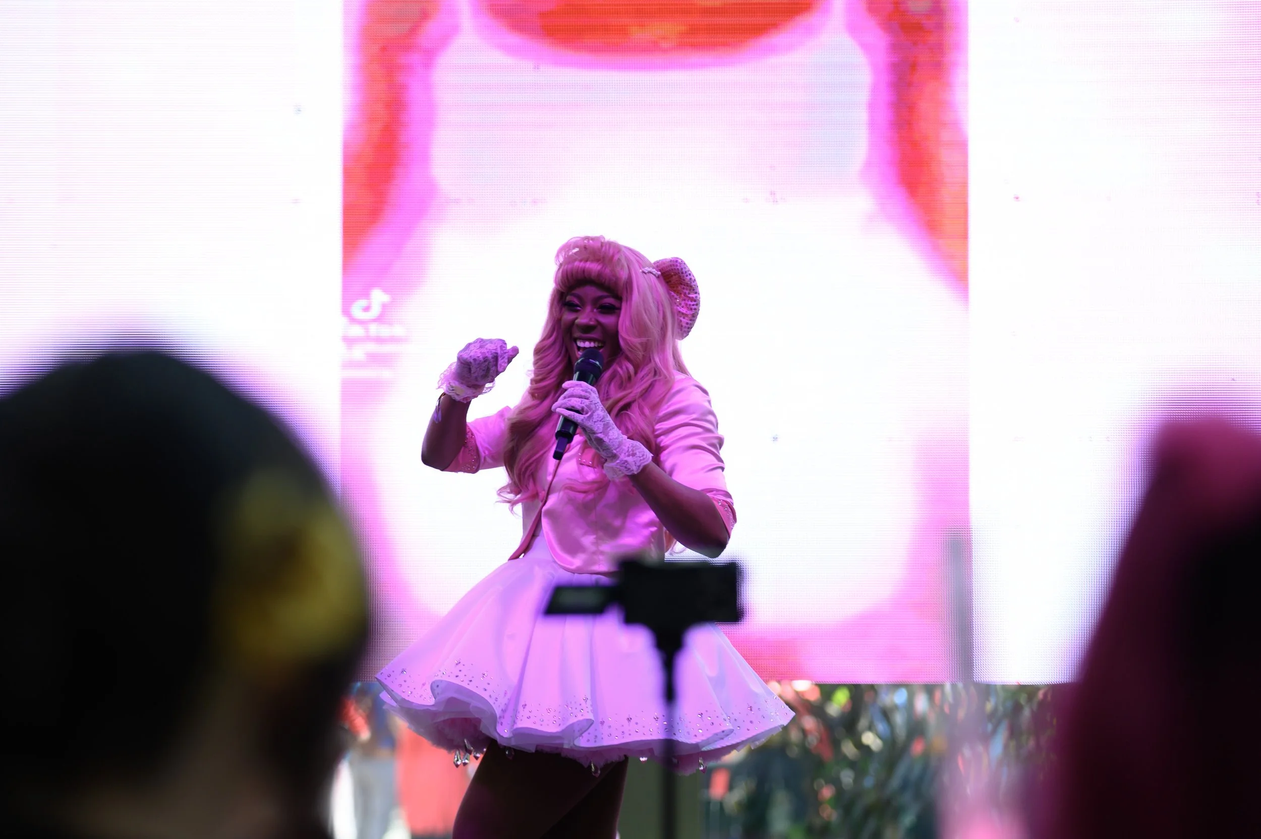 A woman with long pink hair dressed in a pink outfit and gloves, smiling and holding a microphone on stage with a large pink and white digital display in the background.