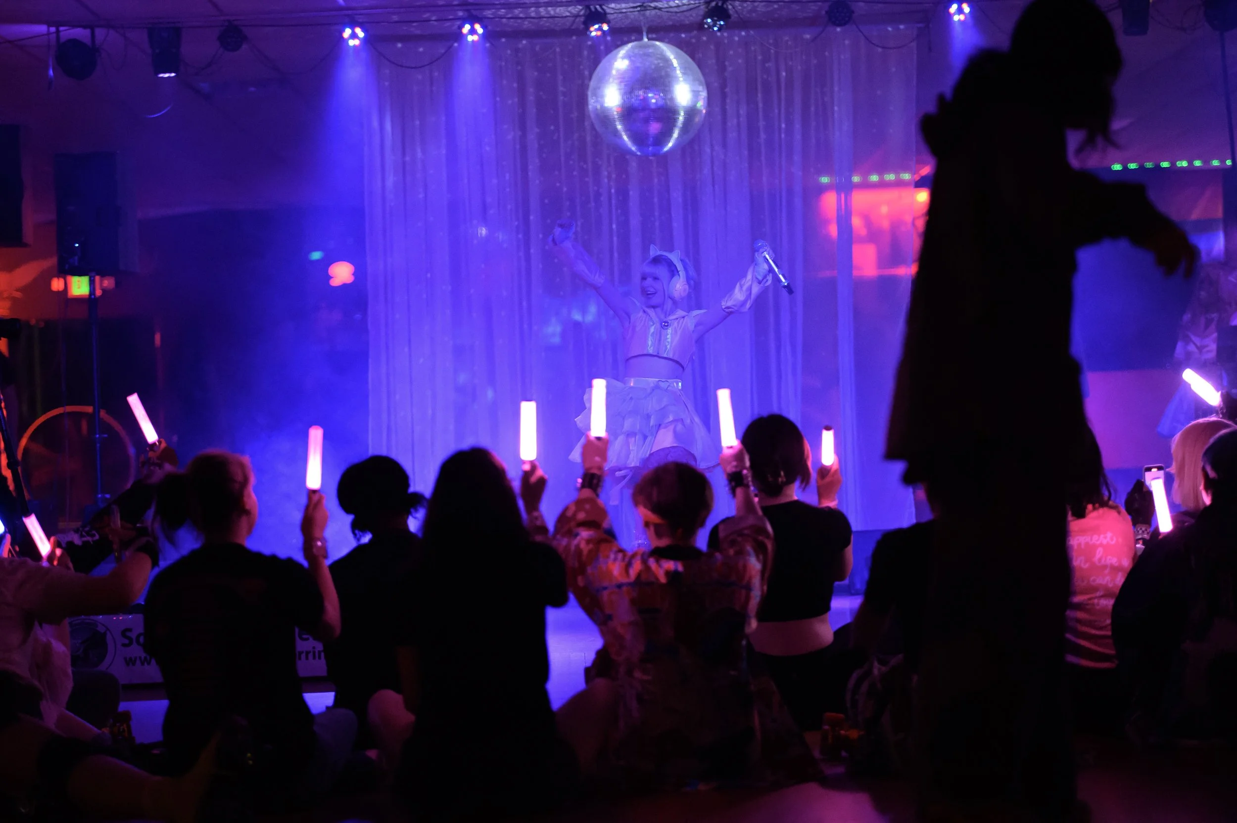 People watching a performer on stage, with some holding pink glow sticks, in a dark, colorful nightlife scene, with a pink costume on the performer and a disco ball overhead.