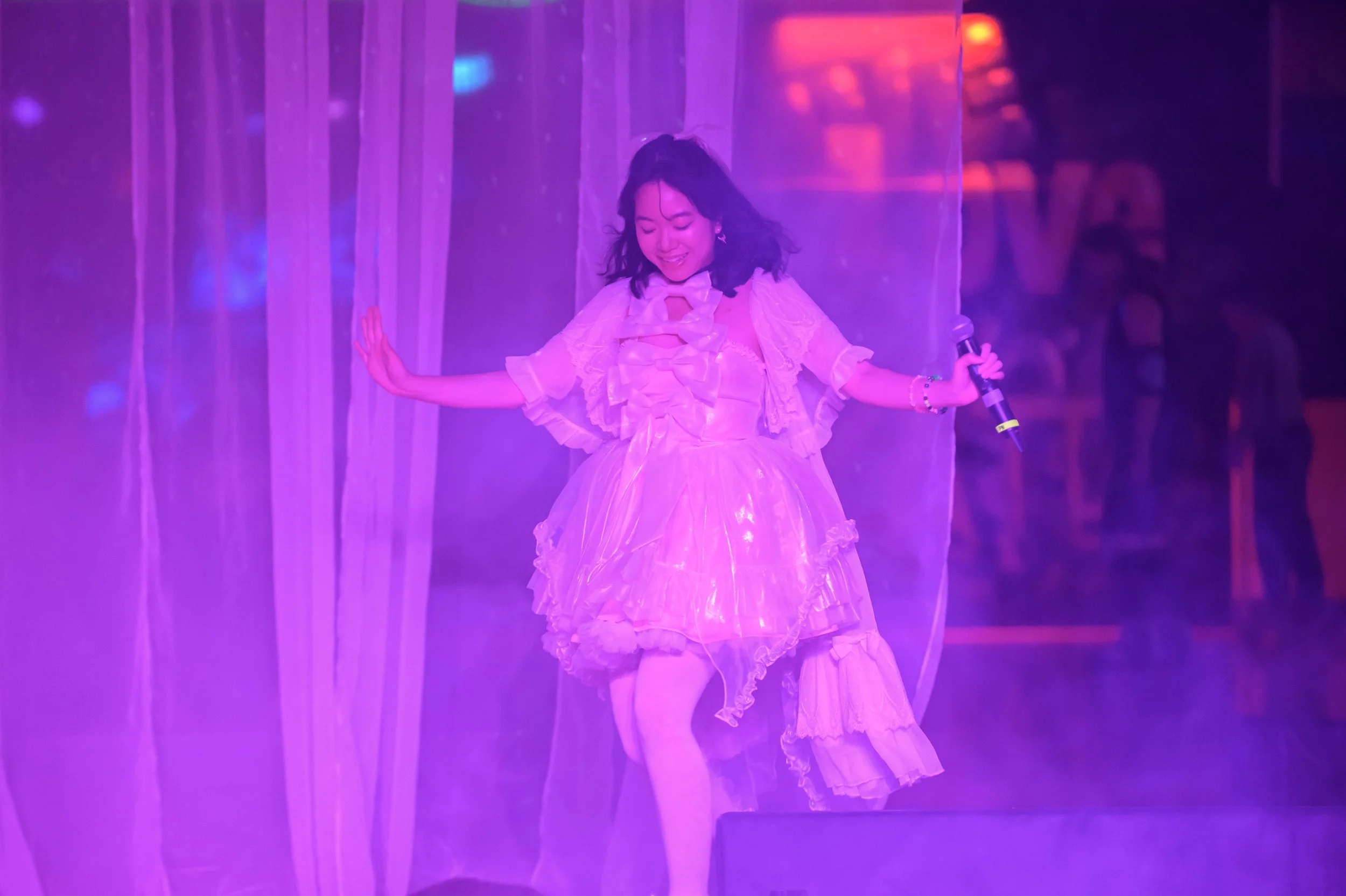 A woman dancing on stage with purple lighting, wearing a lace pink dress, holding a microphone, smiling, and surrounded by sheer curtains.