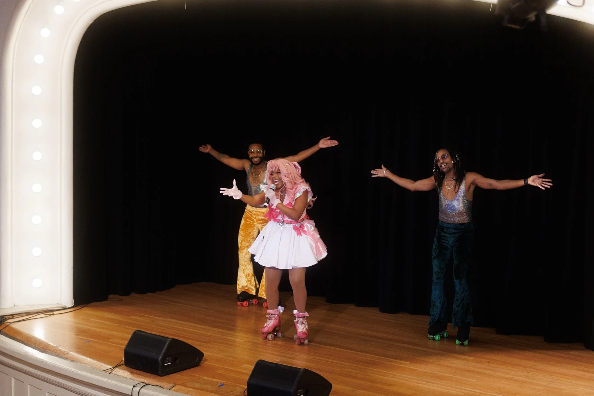 Three performers on a stage with a black curtain background, wearing colorful outfits and roller skates, smiling and dancing.