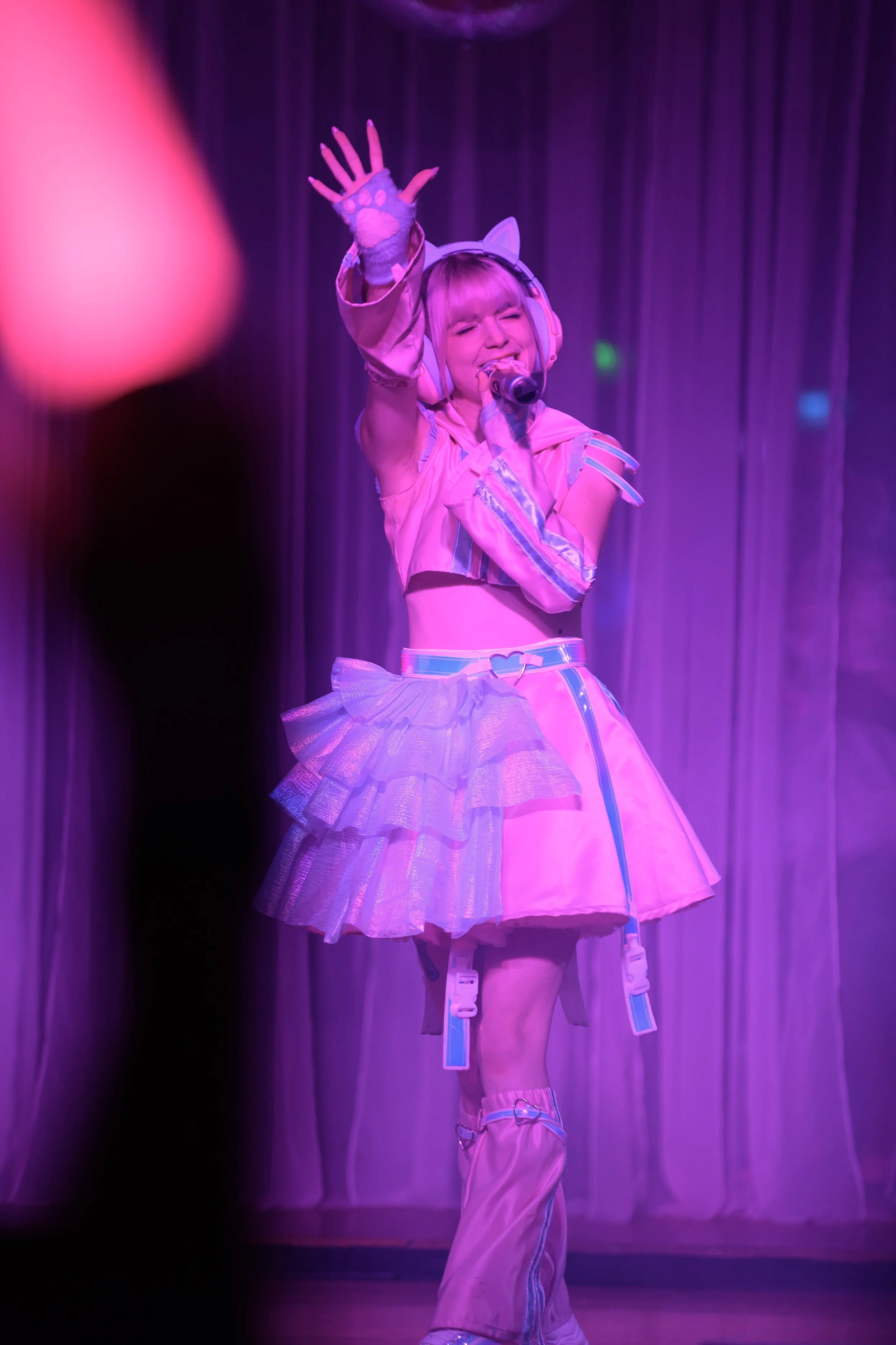 A woman dressed in a pink themed costume with pastel colors, resembling a character or performer, singing into a microphone on stage with purple lighting.