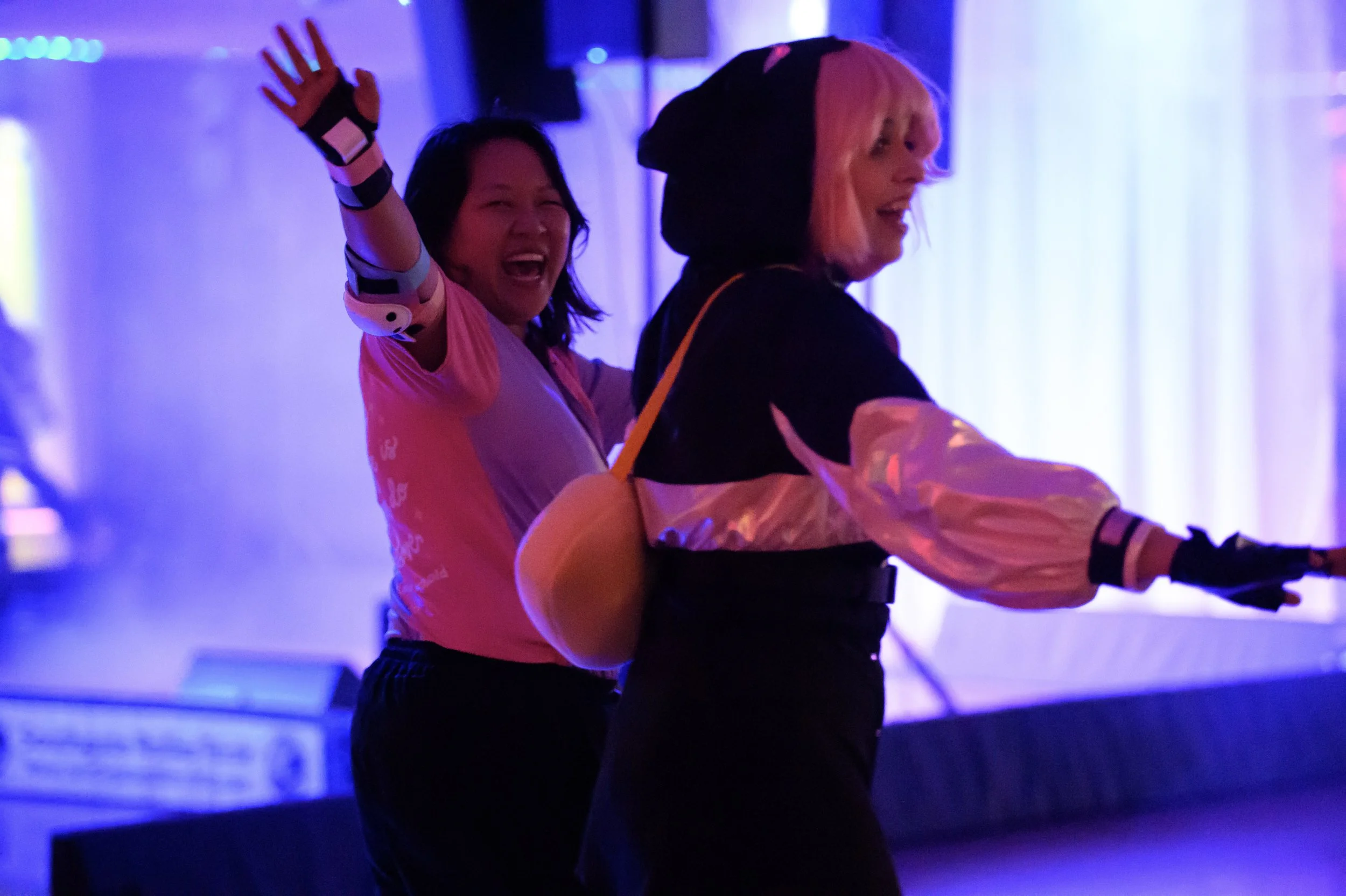 Two women roller skating in a dimly lit room with colorful stage lighting, smiling and having fun.