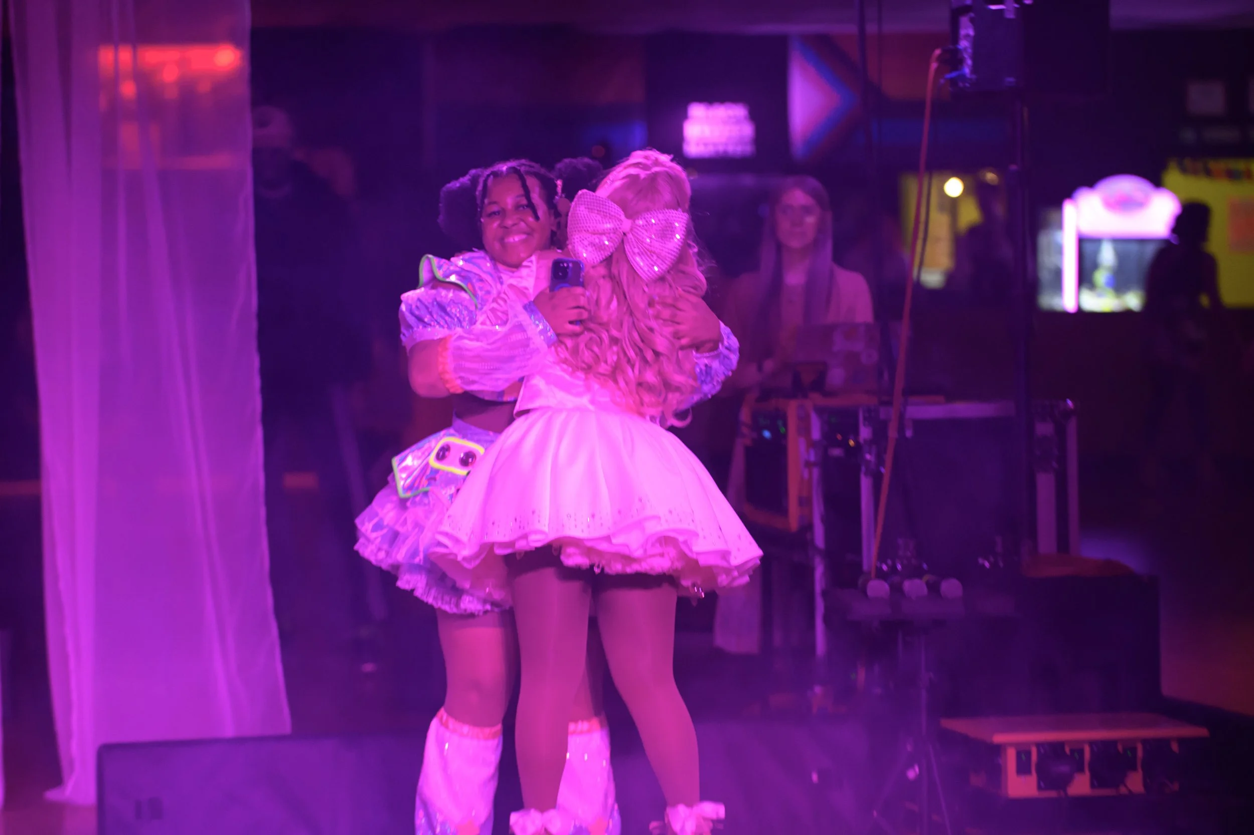 Two women dressed in colorful, playful costumes embracing each other in a dimly lit, neon pink-lit room, one taking a selfie.