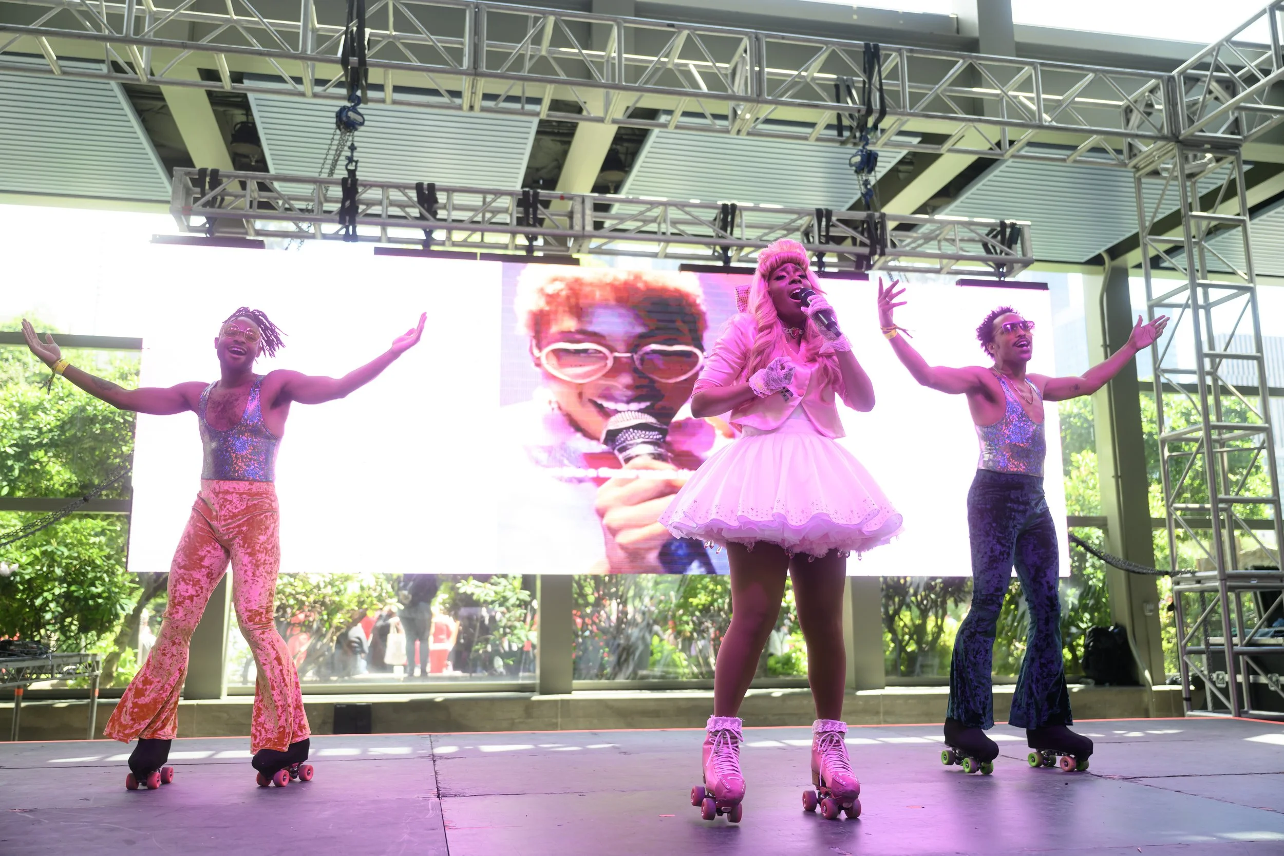 Three performers on roller skates on a stage, with a large screen behind them displaying a smiling person with glasses holding a microphone. The performer in the center is dressed in pink with a tutu and matching pink roller skates, singing into a mi