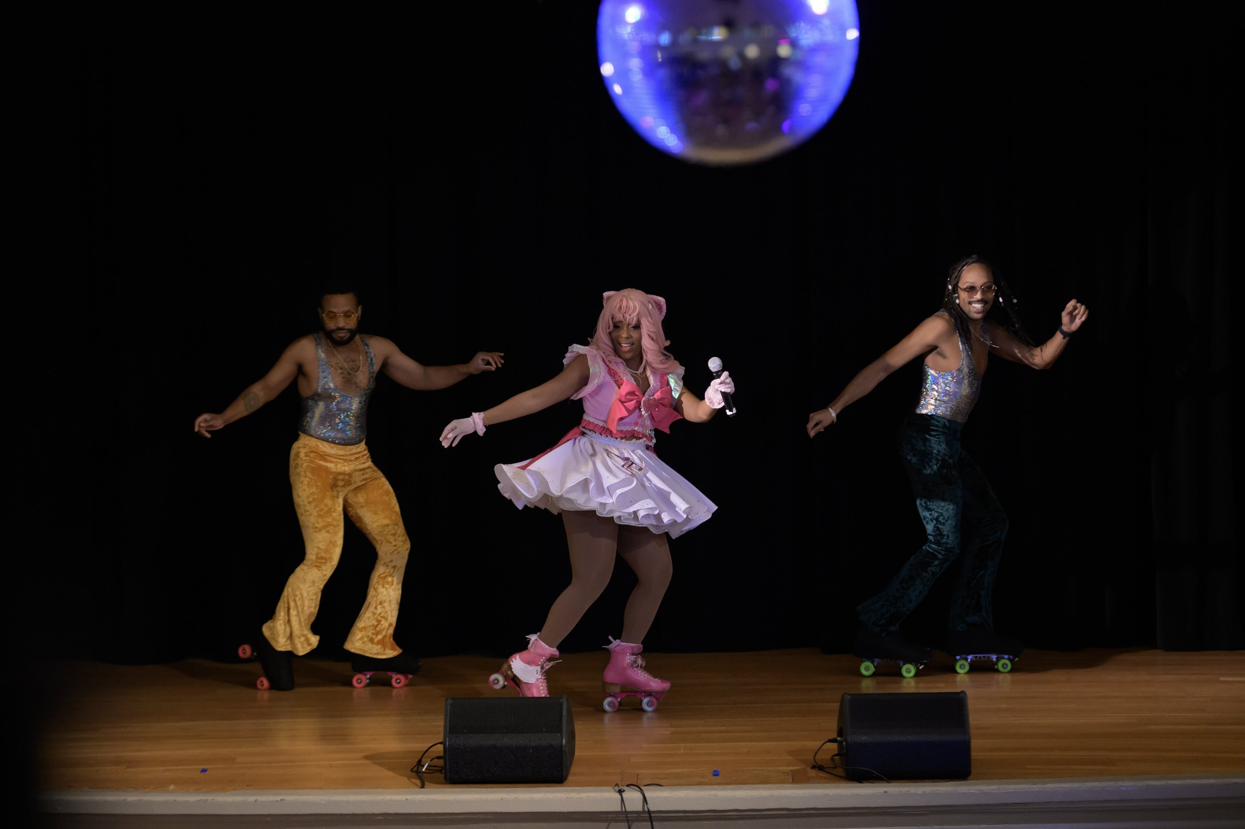 Three performers on stage roller skating, the central figure is dressed as an anime character with pink hair and a pink and white costume, holding a microphone. The stage has a black background with a disco ball overhead.