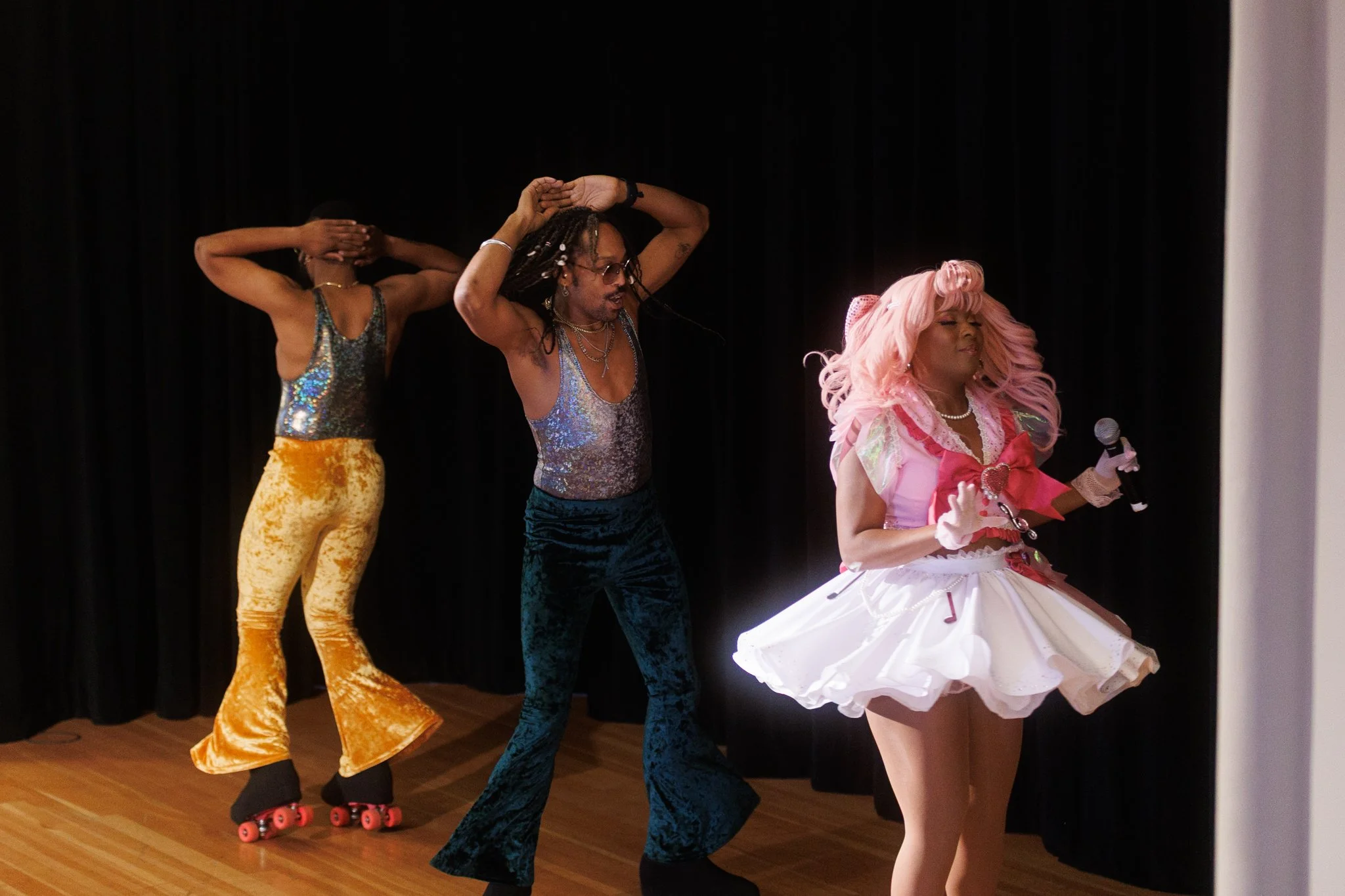 Three performers on stage, two dancing and one singing into a microphone, dressed in colorful outfits with roller skates and glittery clothing, against a black curtain background.