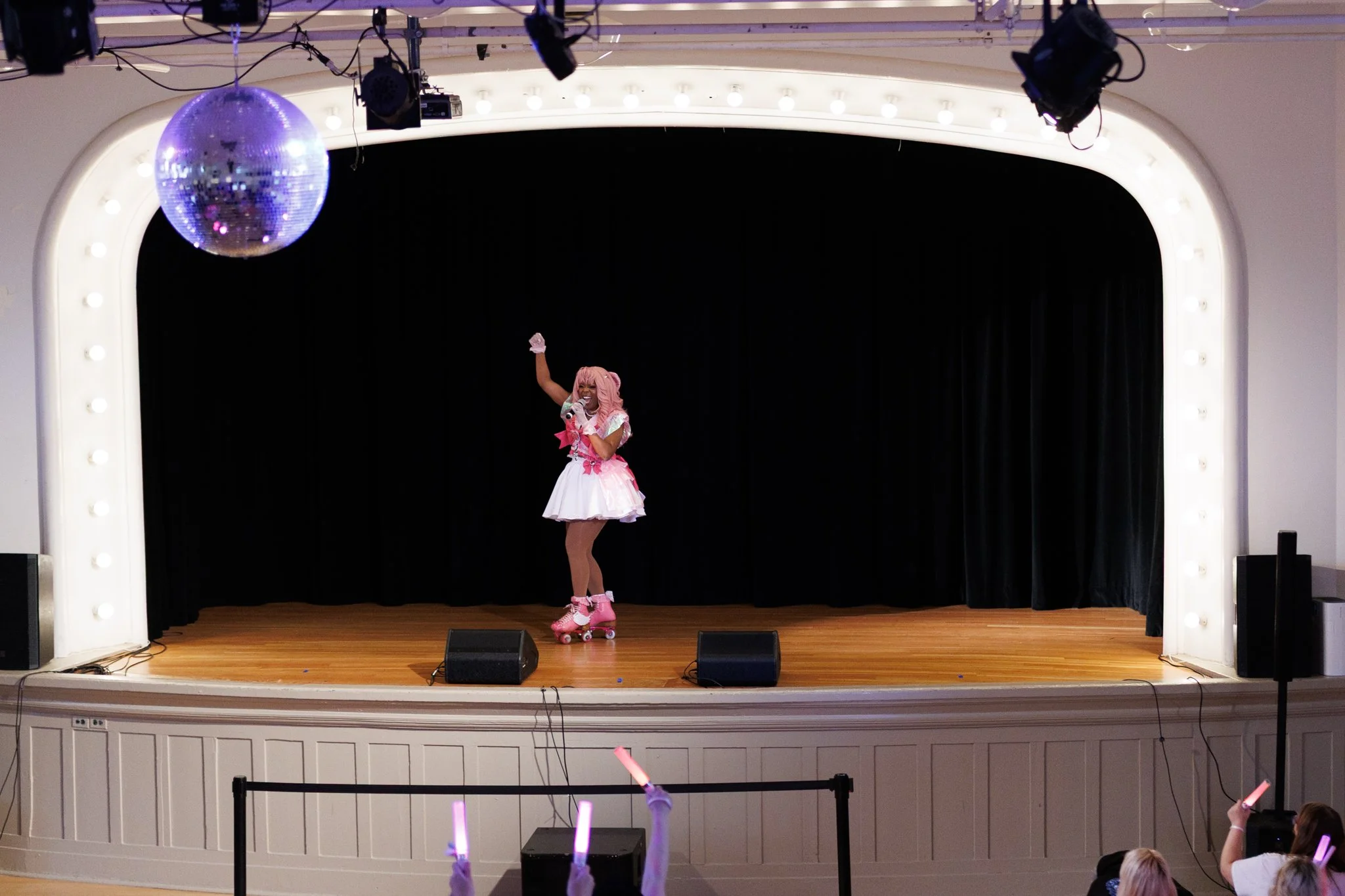 Person dressed in a colorful, animated costume, roller skating on stage, holding a microphone, performing in front of a black curtain, with an audience waving glow sticks nearby.