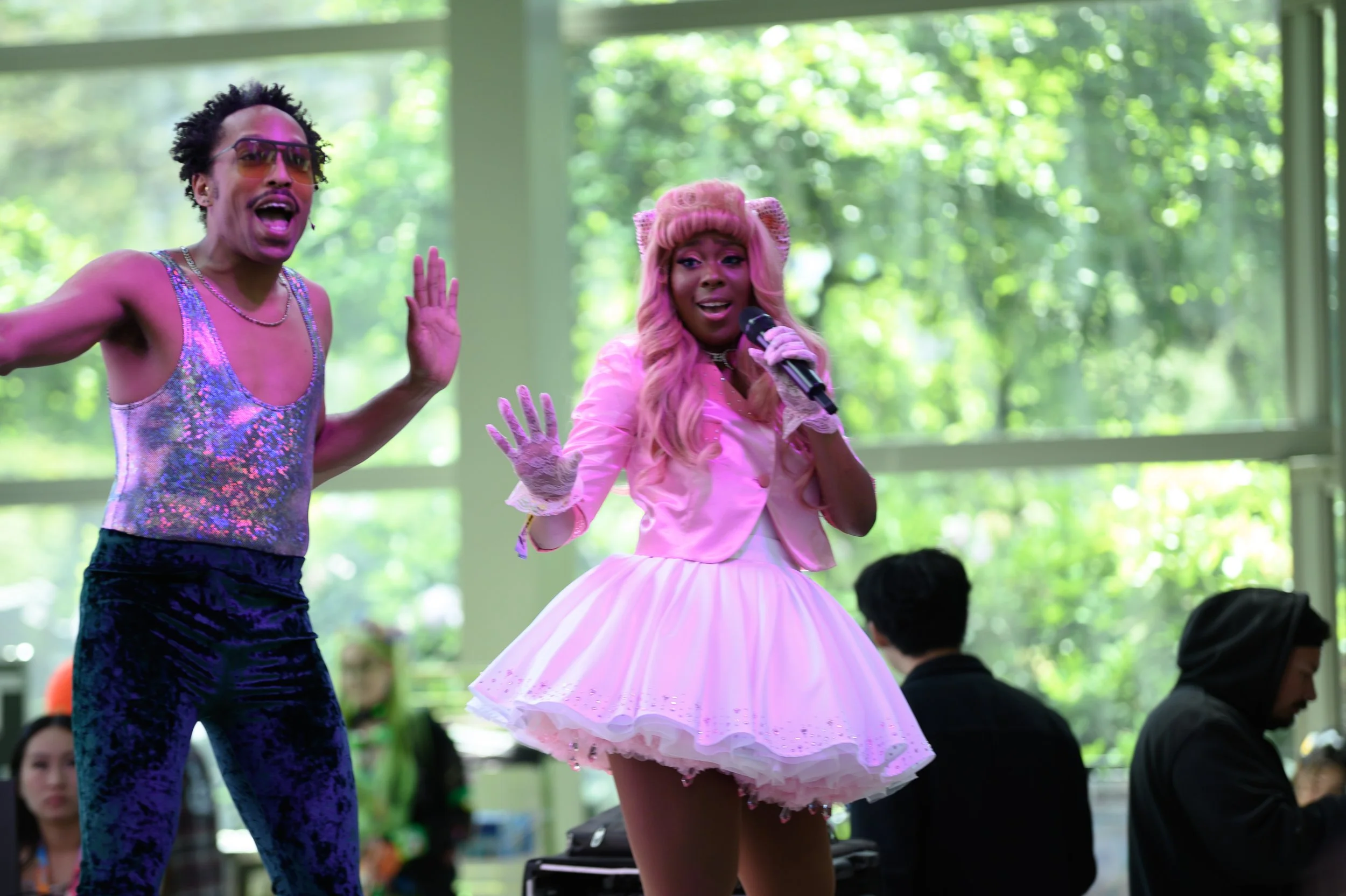 Two performers on stage, one wearing a shiny, sleeveless top, dark patterned pants, and sunglasses, the other dressed in a pink and white dress with a full skirt, pink gloves, and pink hair, singing into a microphone.