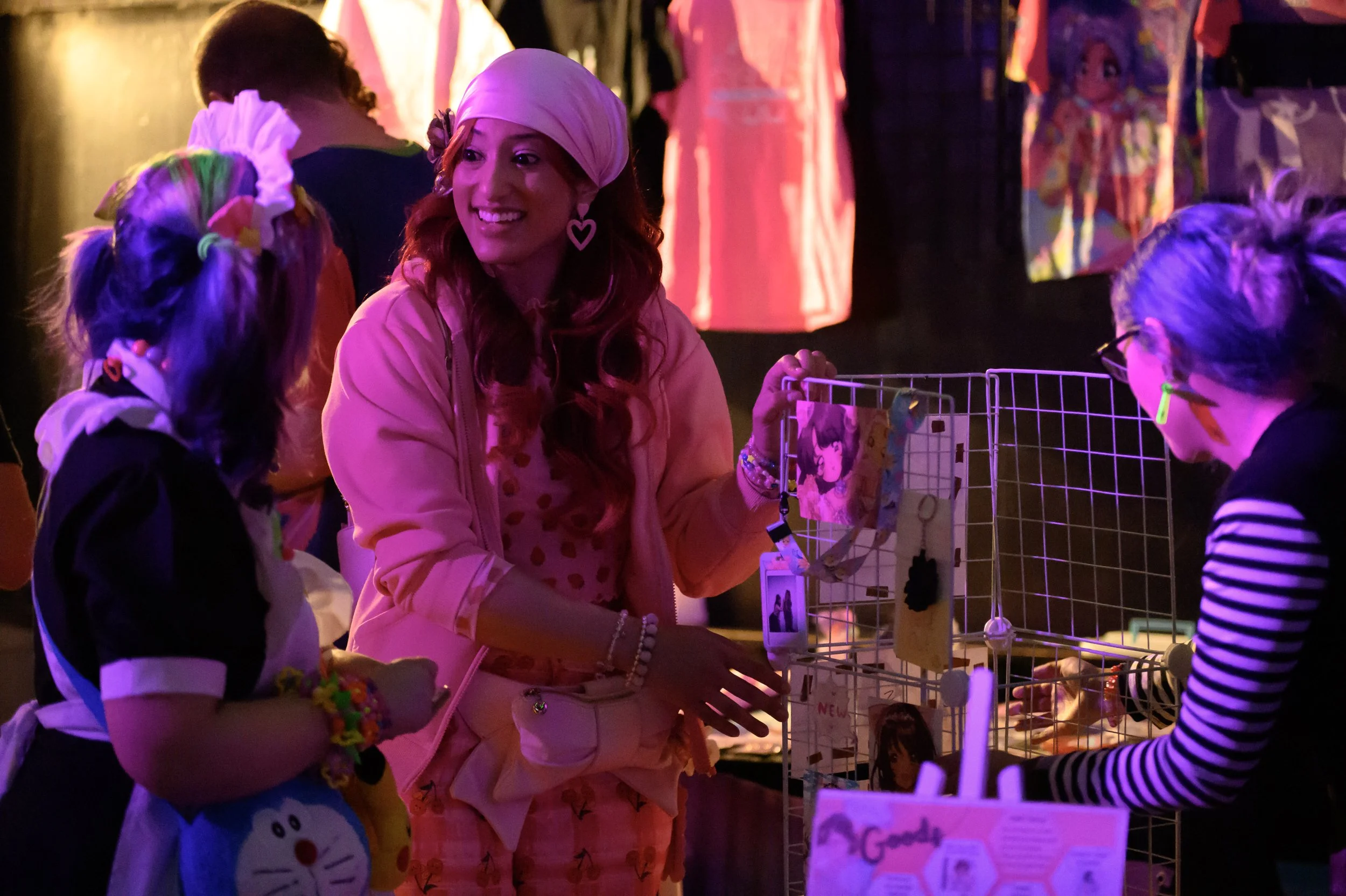 A woman with long red hair wearing pink pajamas and a pink headscarf talking to a girl with glasses and colorful hair at a booth. The booth has photos and items hanging on a wire grid. The scene is lit with pink and purple lights.