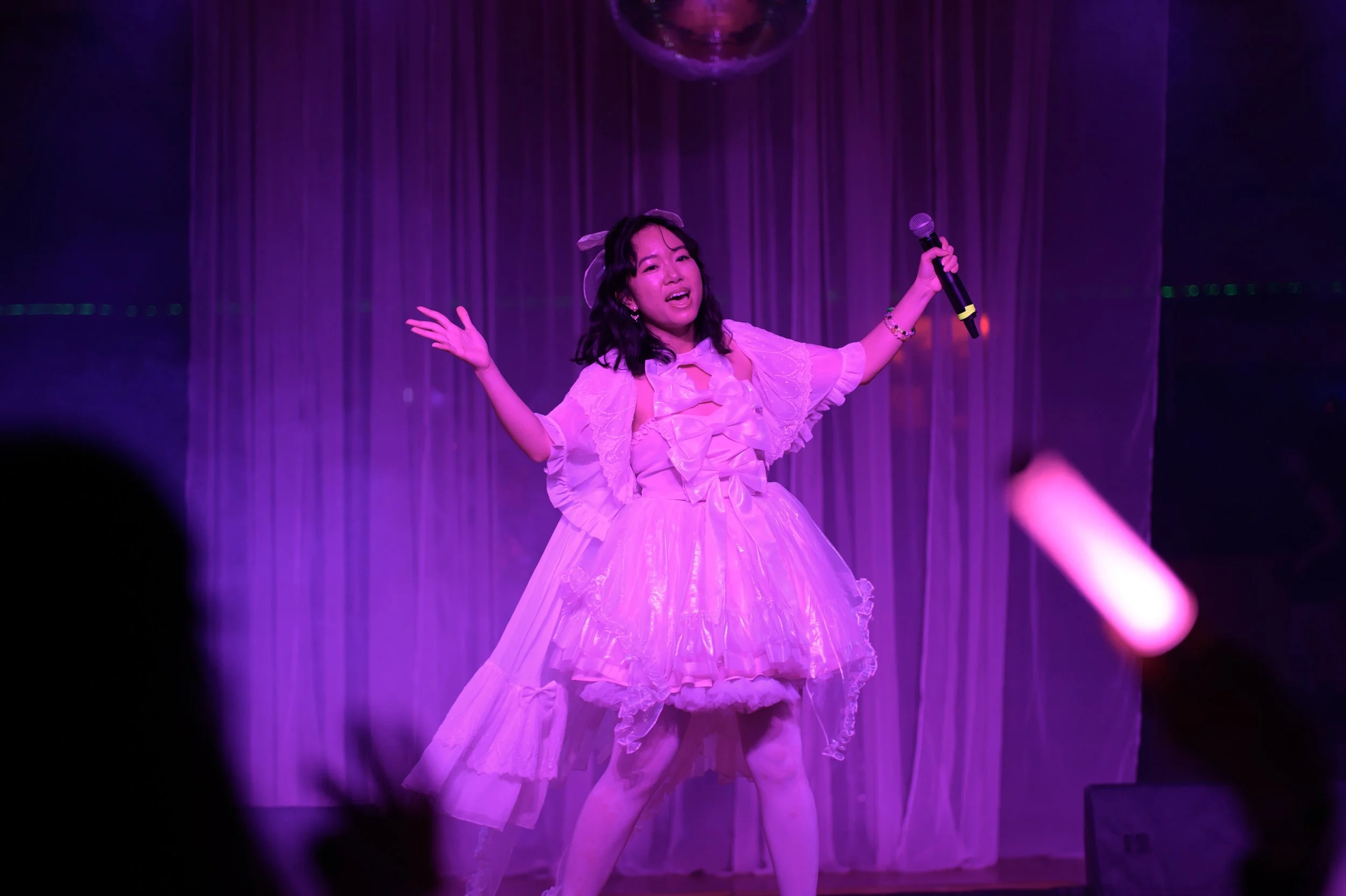 A young woman on stage singing into a microphone, wearing a pink, ruffled dress with puffed sleeves and a matching bow in her hair, with purple lighting and curtains in the background.