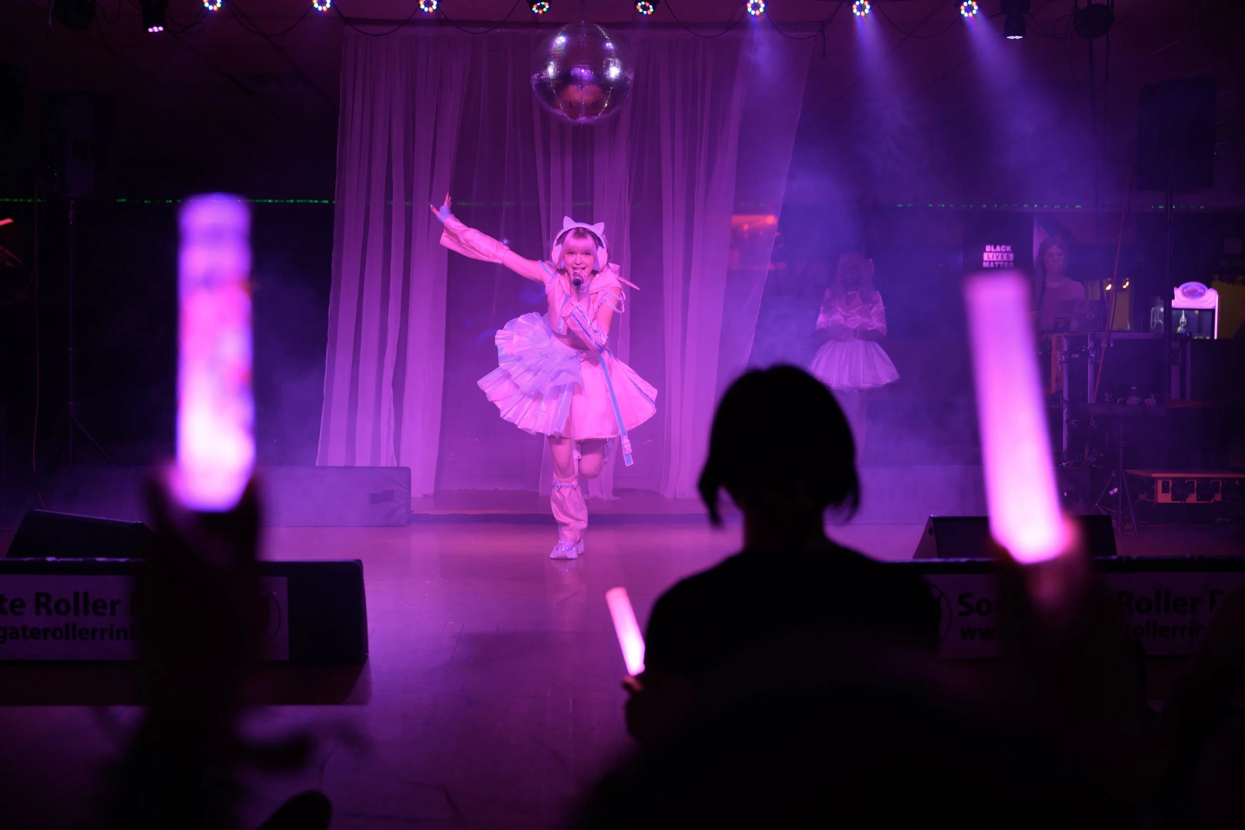 Performer in a pink ballet costume on stage with purple lighting, a disco ball overhead, and people in the audience holding glowing pink sticks.