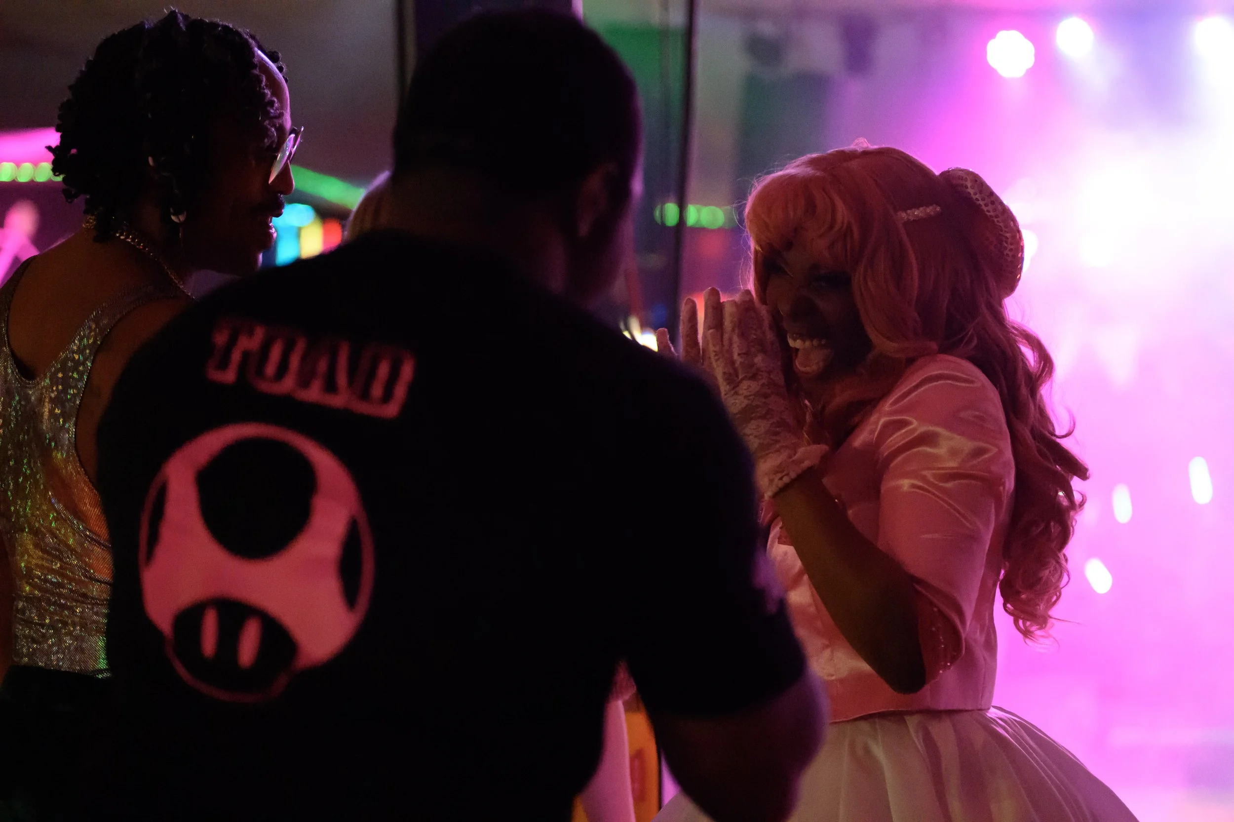 People in colorful costumes at a party or event, illuminated by pink and purple lights.