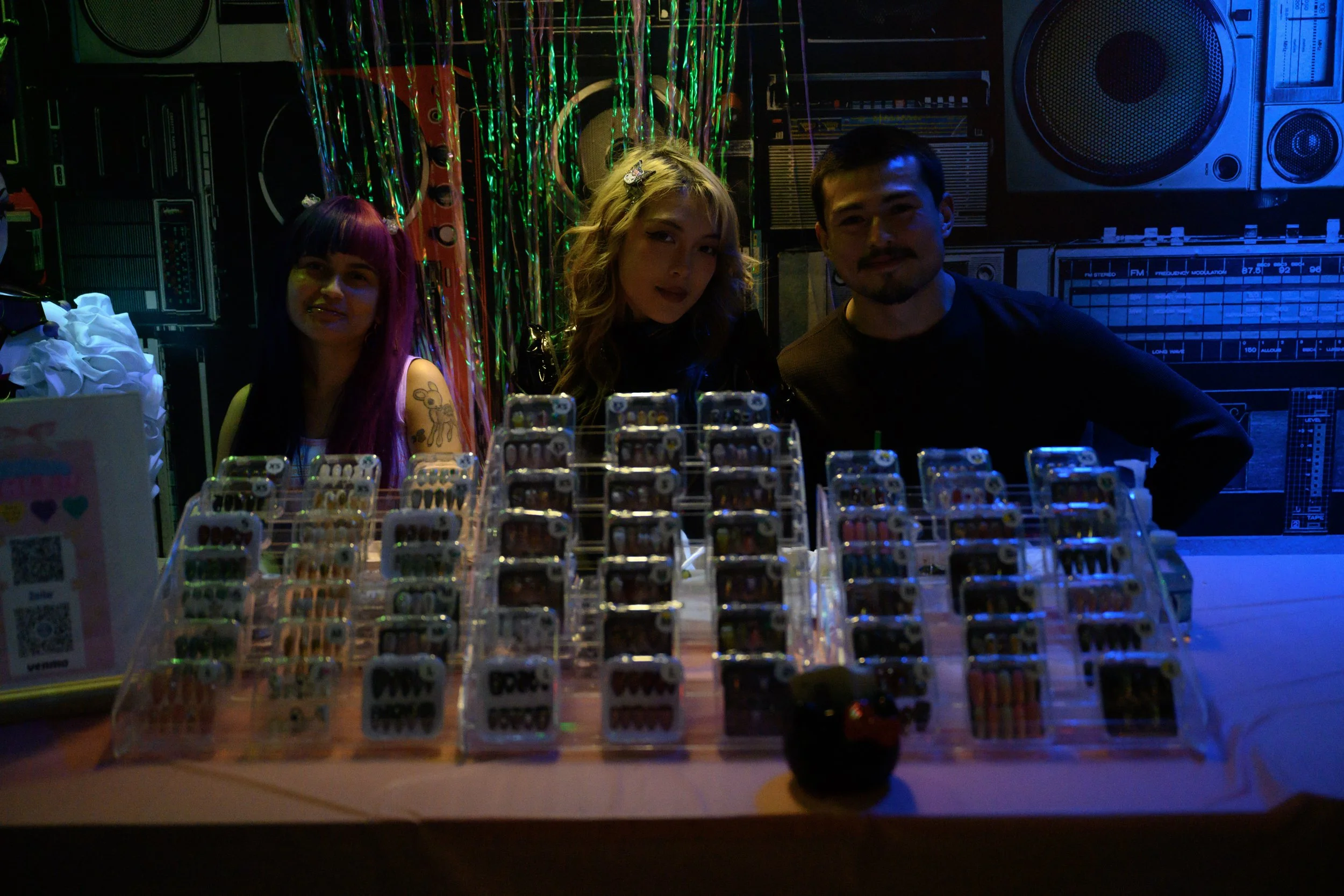 Three people sitting behind a table with candies in display cases at a party. The background has colorful streamers and equipment, with a blue lighting ambiance.