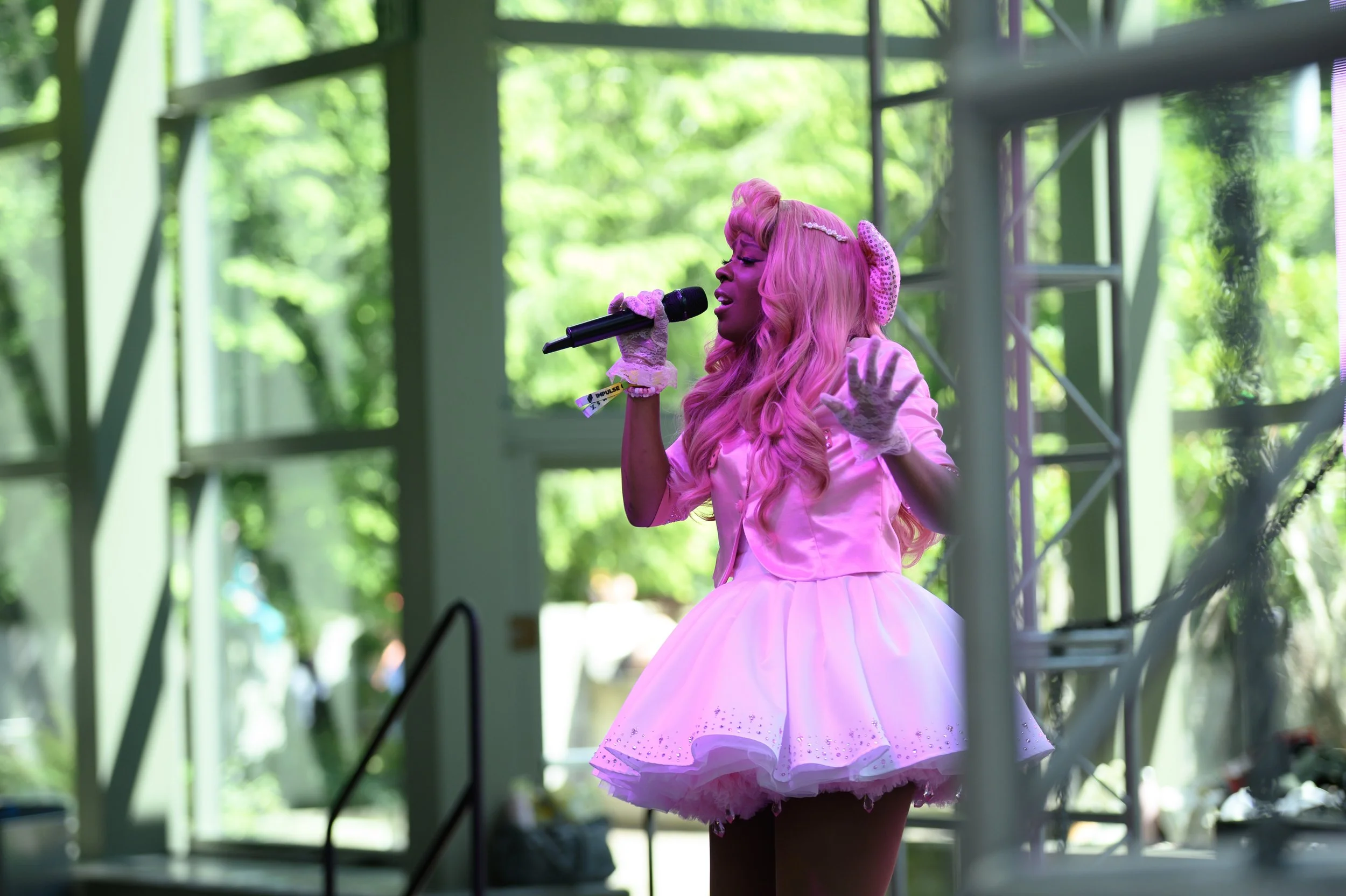 A woman with long pink hair wearing a pink dress and gloves sings into a microphone on stage with a background of green trees outside.