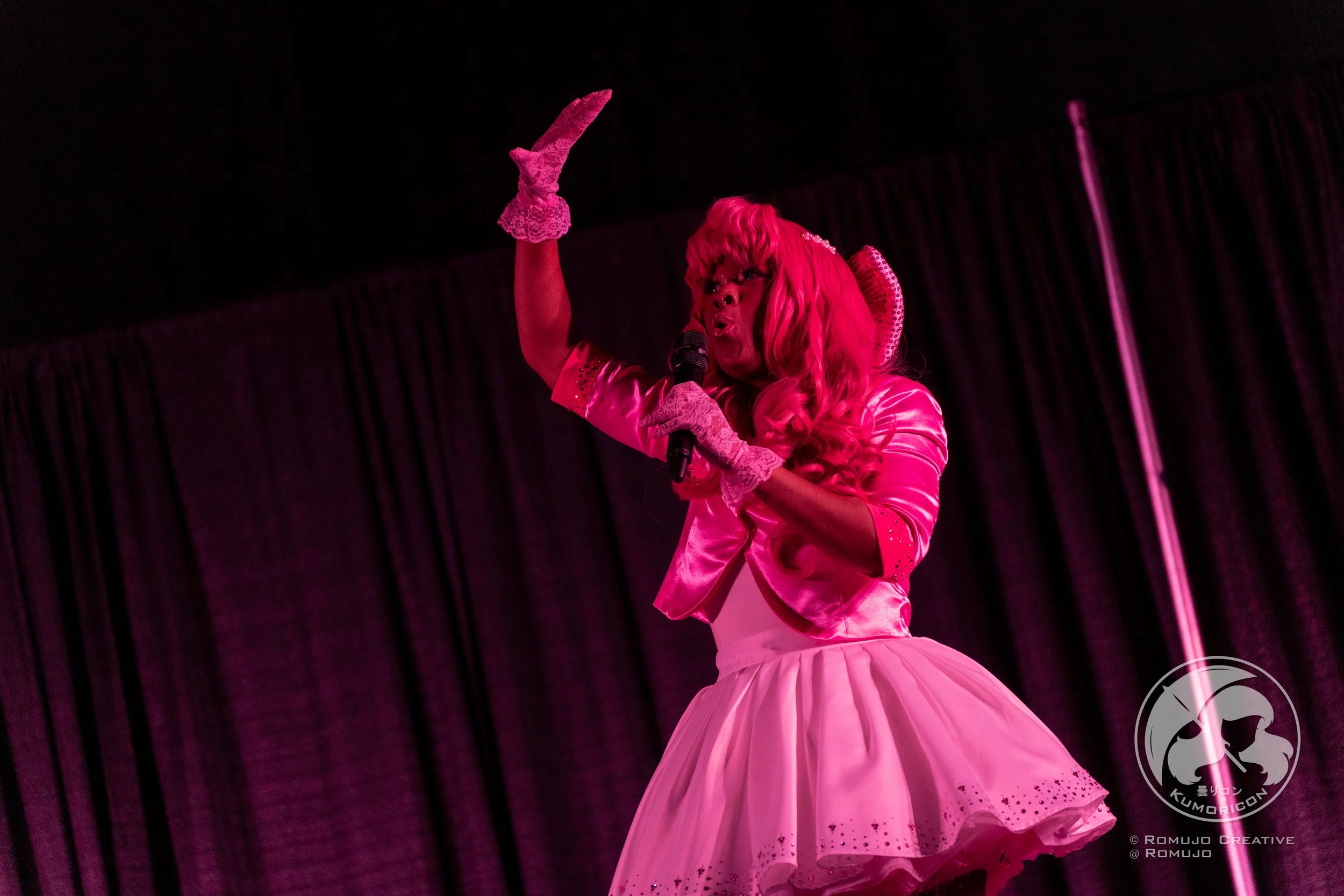 Performer with pink hair and costume singing into a microphone on stage with black curtains.