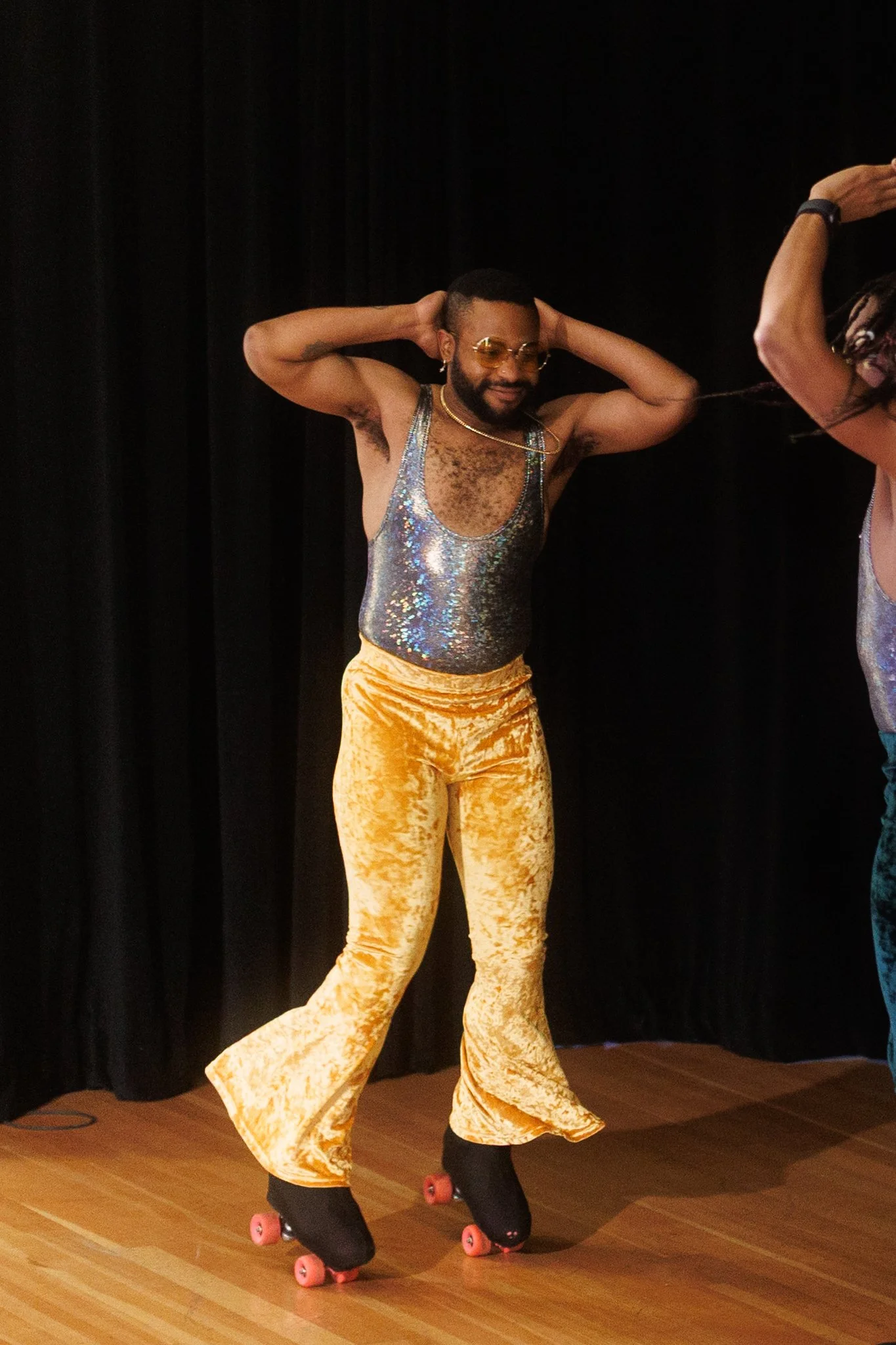 A man wearing yellow velvet pants, a silver shimmering tank top, and roller skates standing on wooden floor with black curtains in the background.