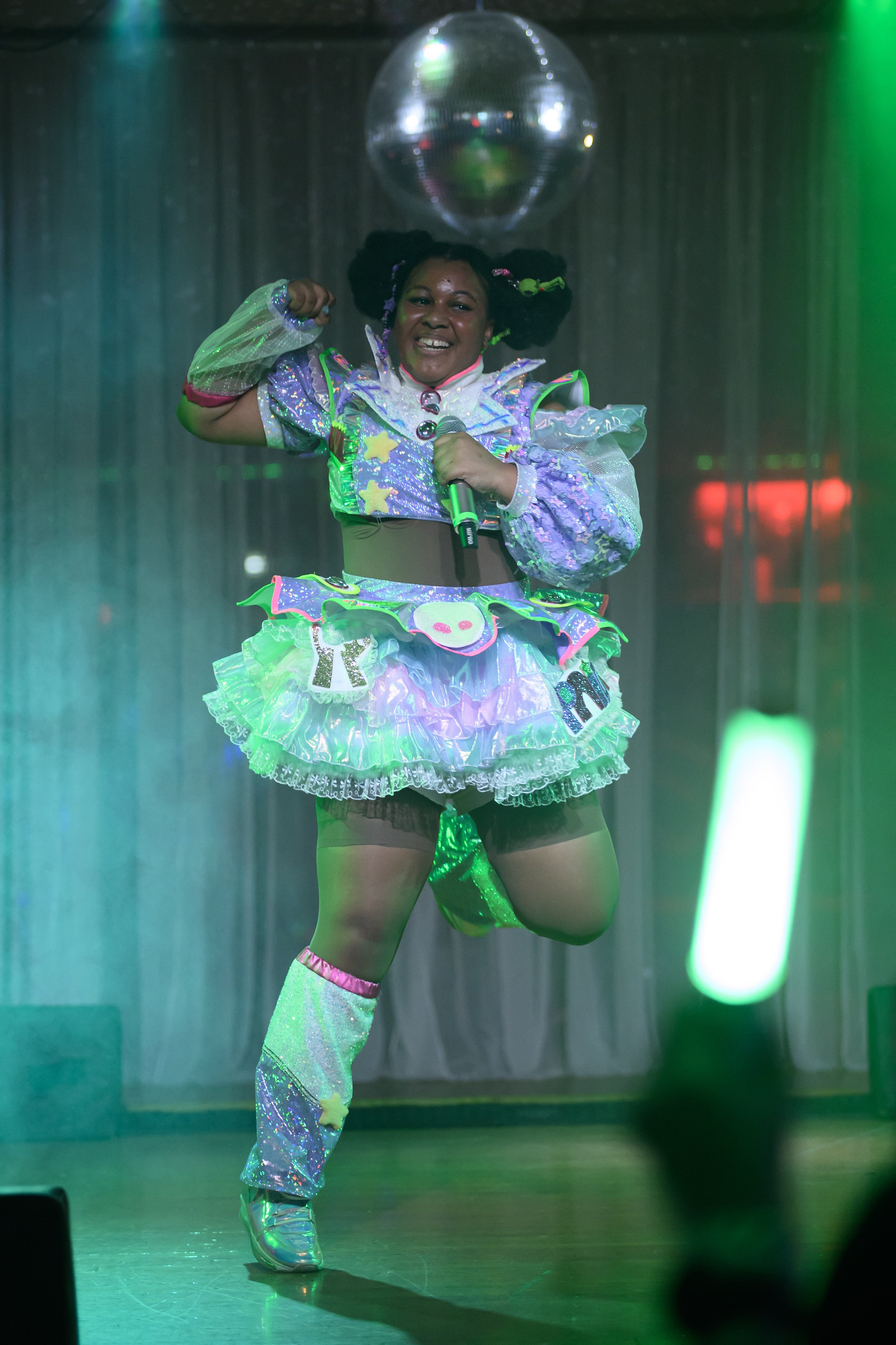 A woman wearing a colorful, shiny, and whimsical costume dances on stage under green lighting, holding a microphone, with a disco ball overhead.