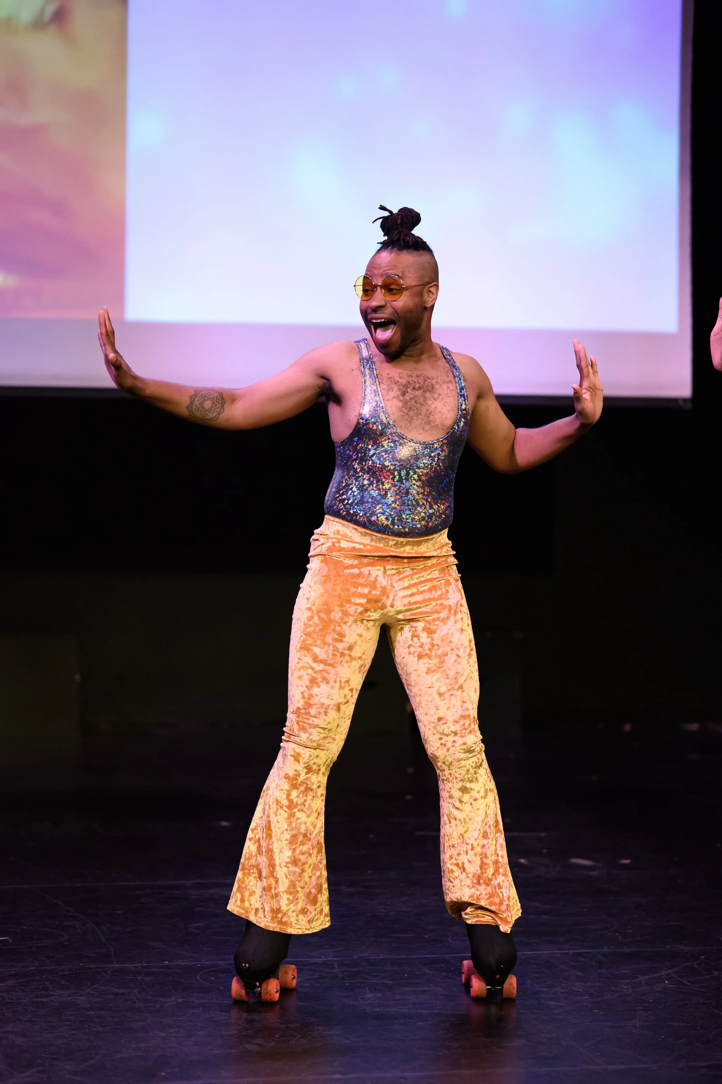 Person with short hair, sunglasses, wearing a sparkly sleeveless top, gold crushed velvet pants, and roller skates, performing on stage with arms outstretched and an enthusiastic expression.