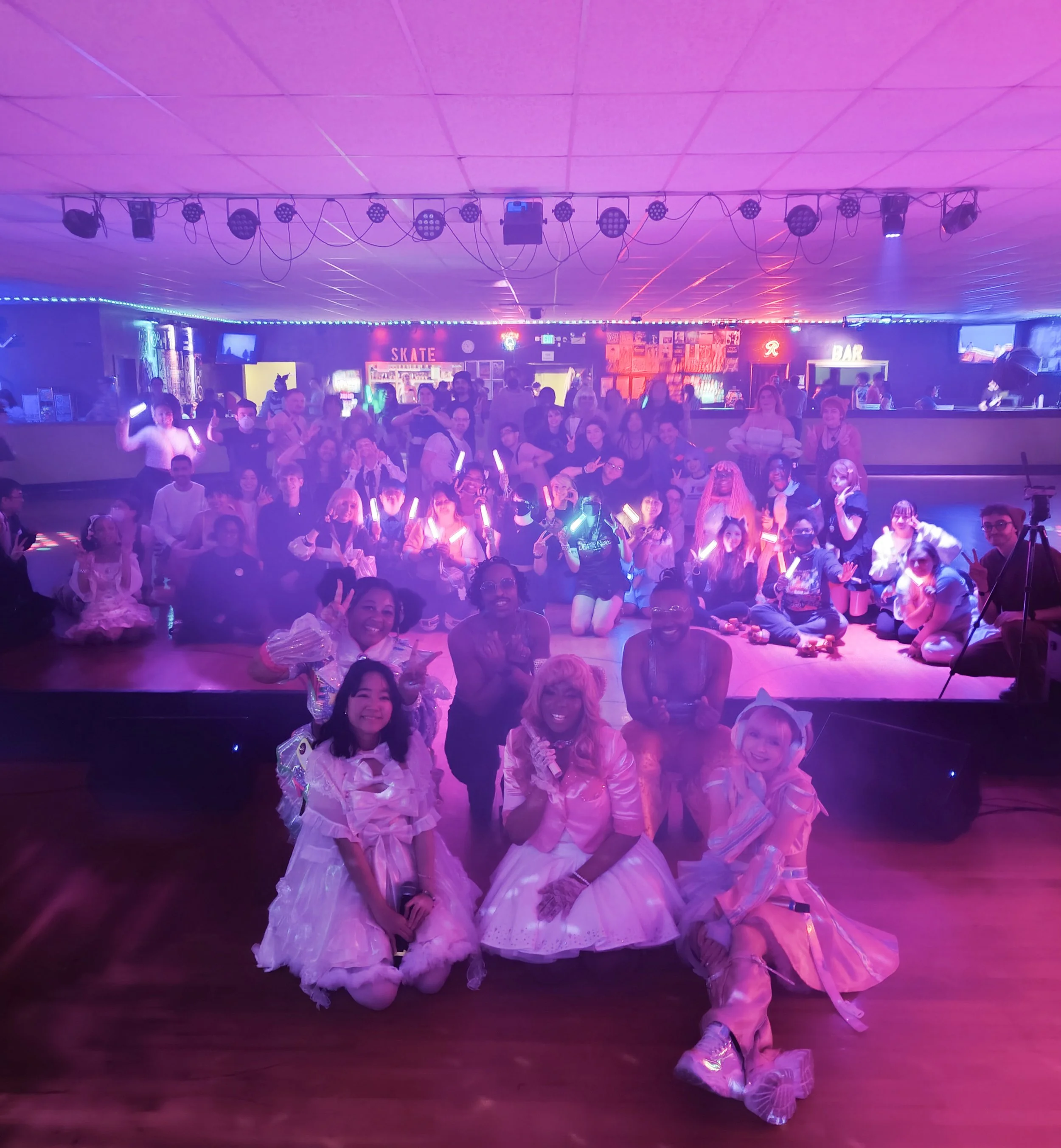 Group photo of people at a roller skating party with glow sticks in a roller rink with neon signs and colorful lighting.